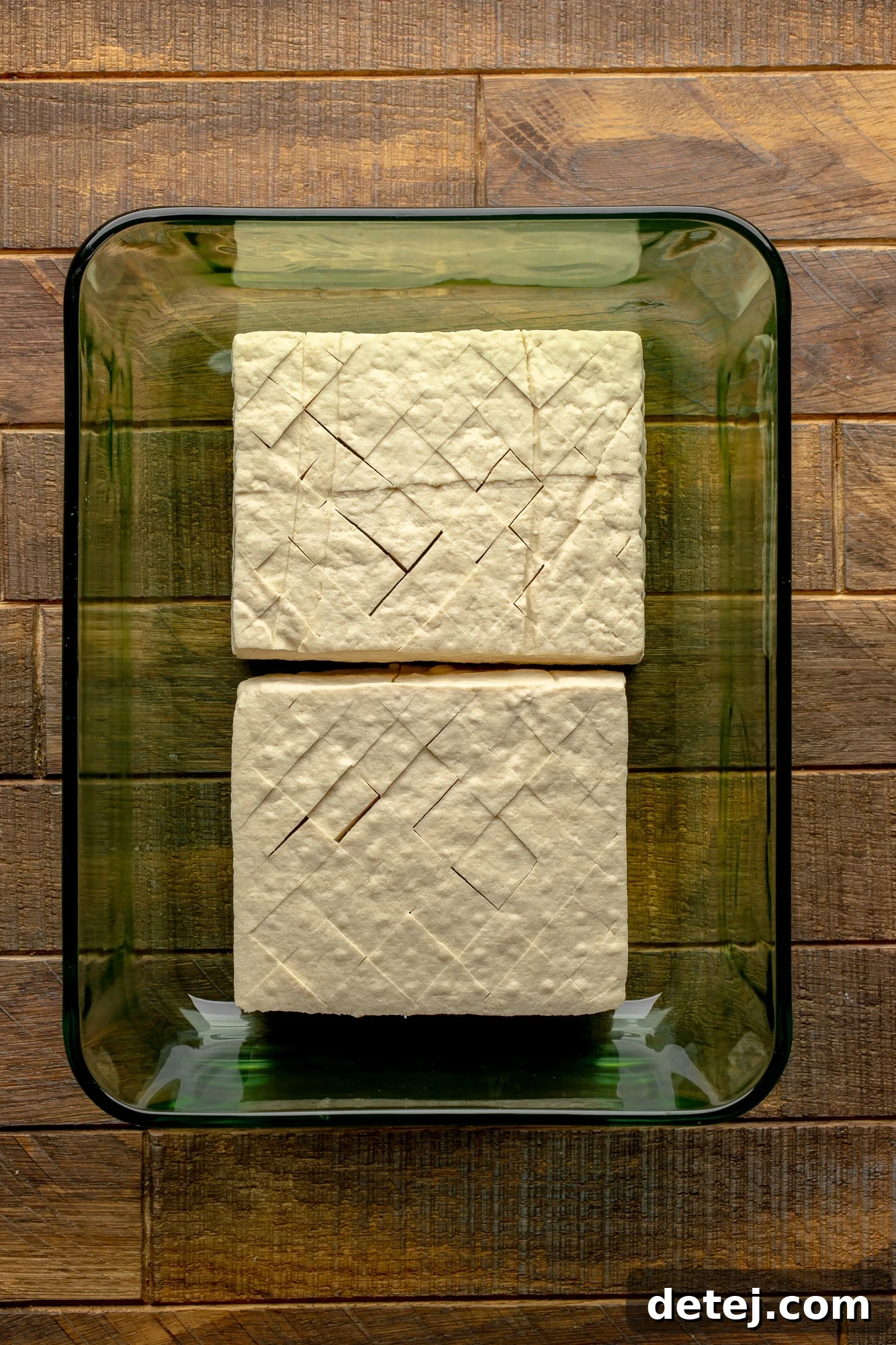 overhead view of 2 scored tofu blocks in a glass container.