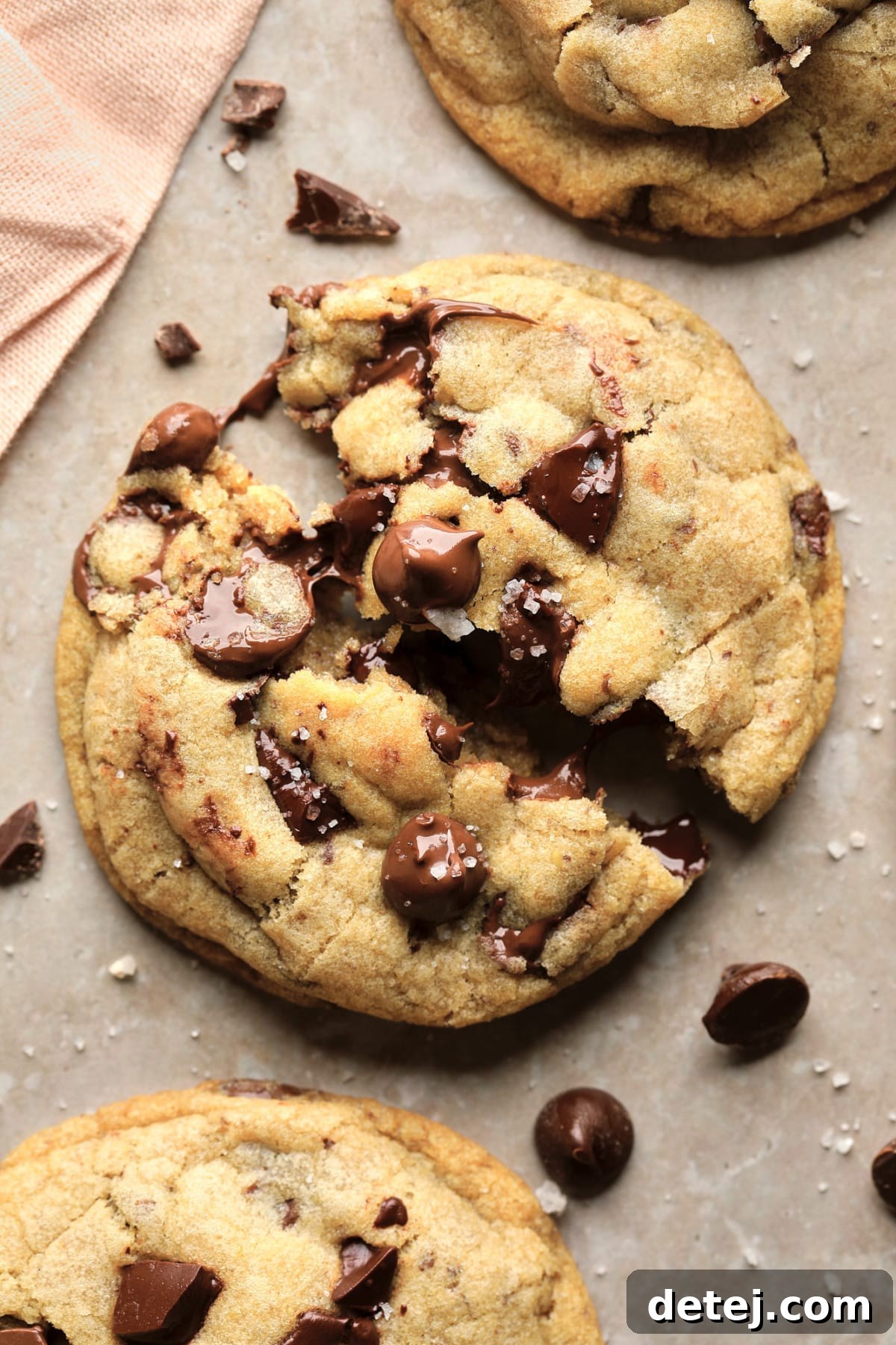 A perfectly broken open vegan chocolate chip cookie, showcasing its soft interior and melted chocolate, resting on a rustic beige background with a sprinkle of coarse sea salt.