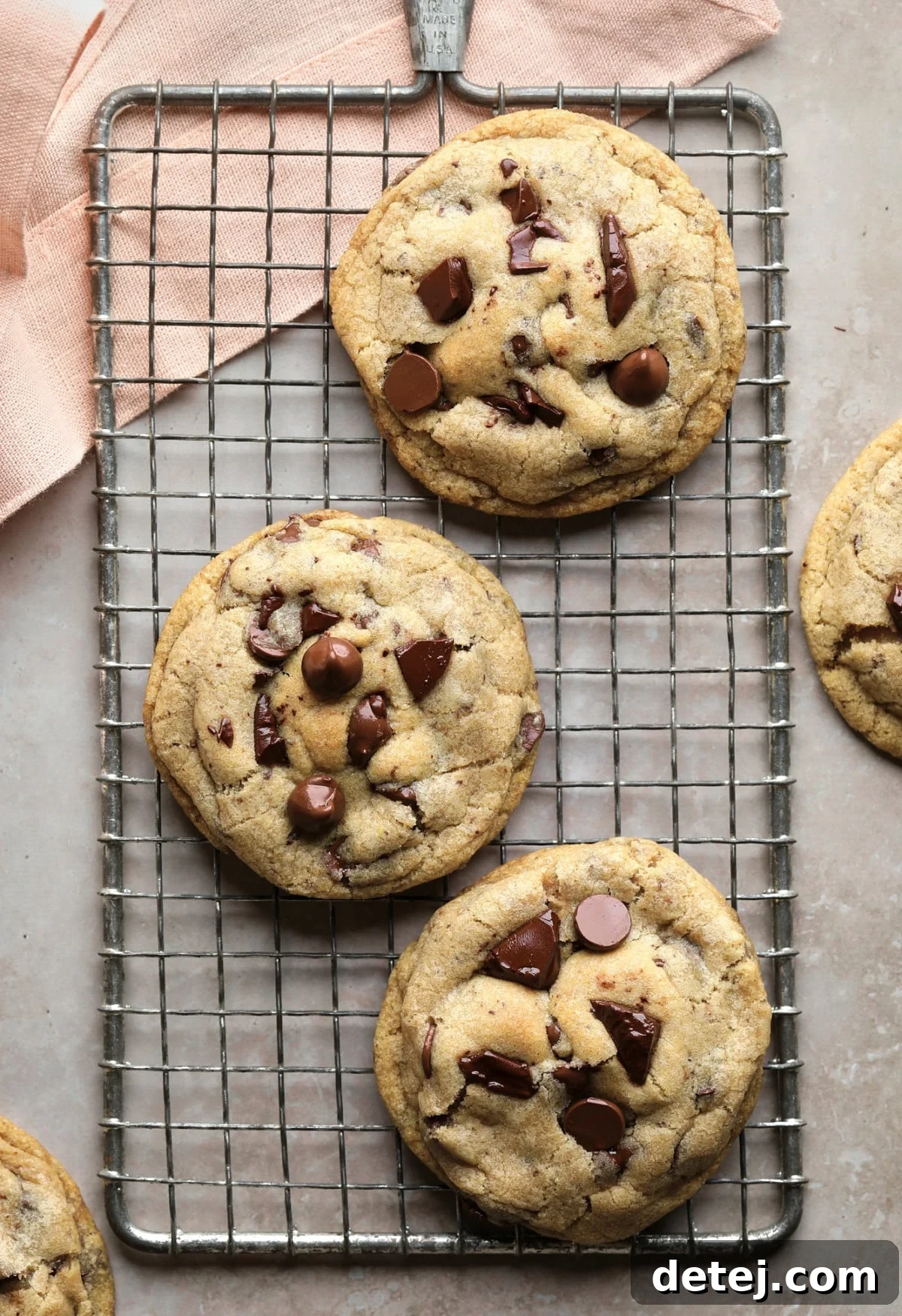 A serene image of a few freshly baked vegan chocolate chip cookies resting on a wire cooling rack, with a hint of a pink towel and a soft, greyish background, signifying a moment of delicious anticipation.