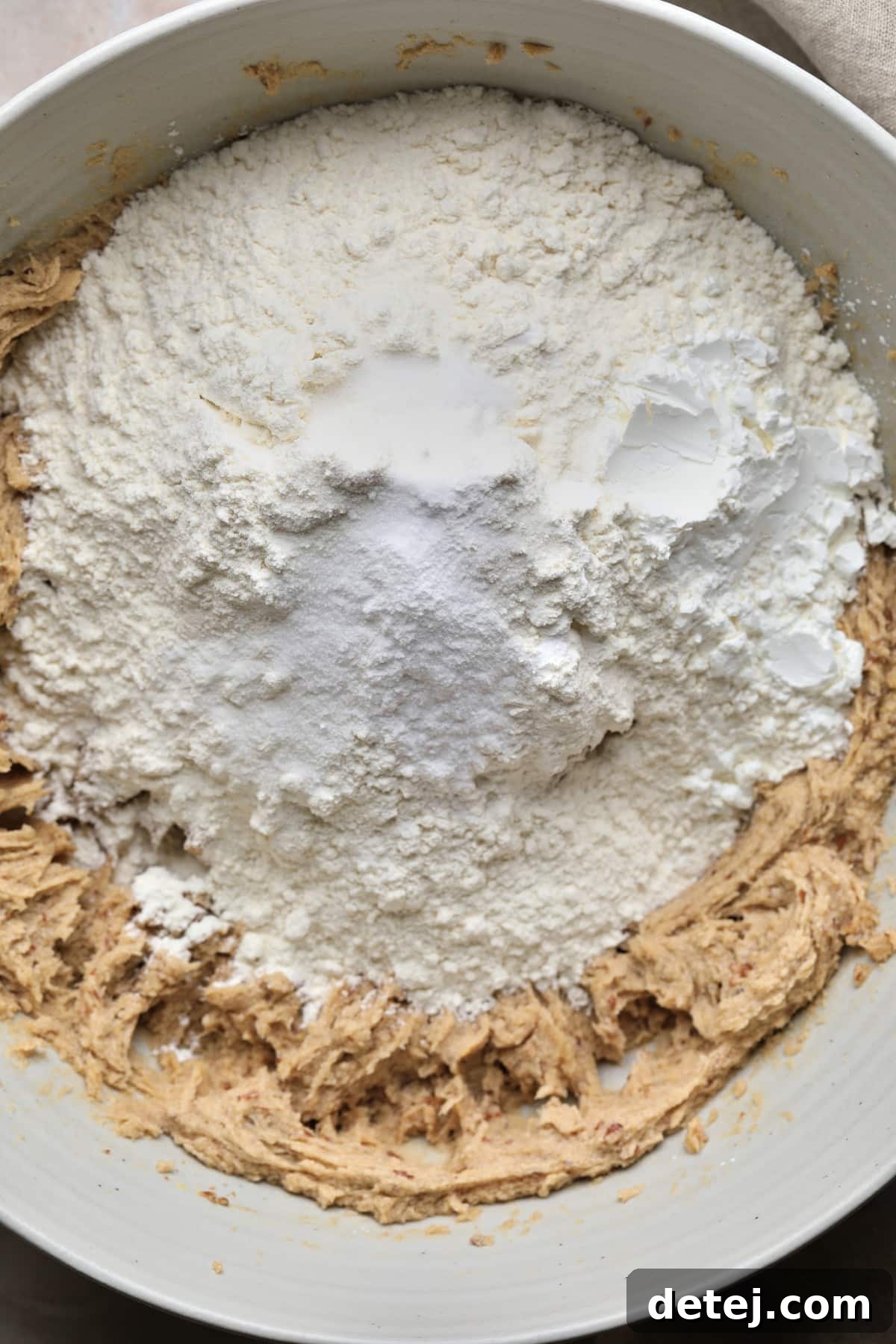 Dry ingredients, including flour, cornstarch, baking soda, and salt, being carefully added on top of the beige wet mixture in the mixing bowl.