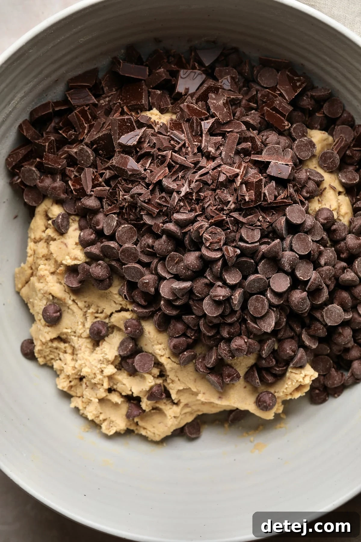 A close-up of delicious vegan chocolate chips and larger chocolate chunks generously piled on top of the ready-to-mix cookie batter in a bowl.