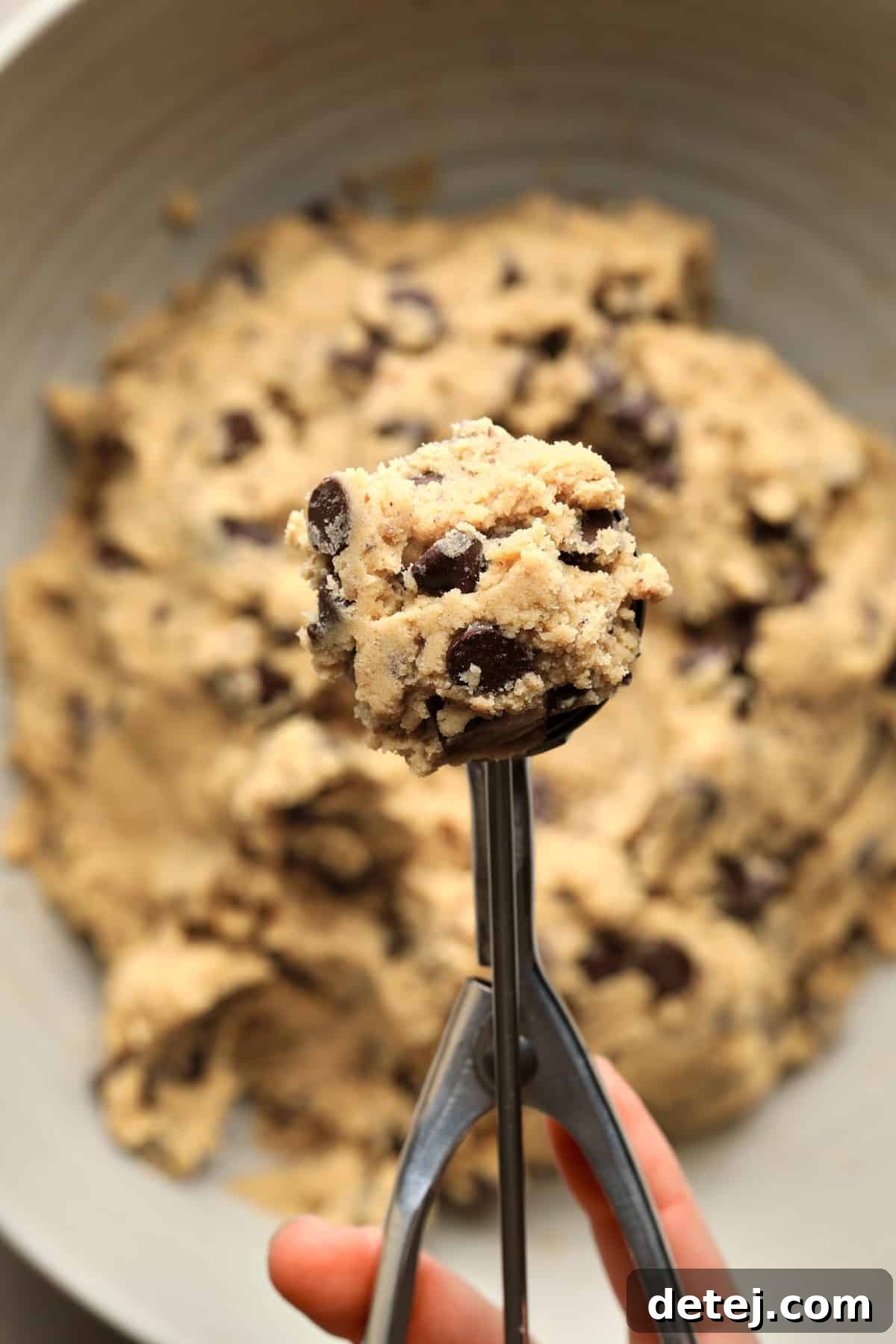 A hand using a cookie scoop to portion out perfect balls of vegan chocolate chip cookie dough from a large bowl, ensuring uniform size for baking.