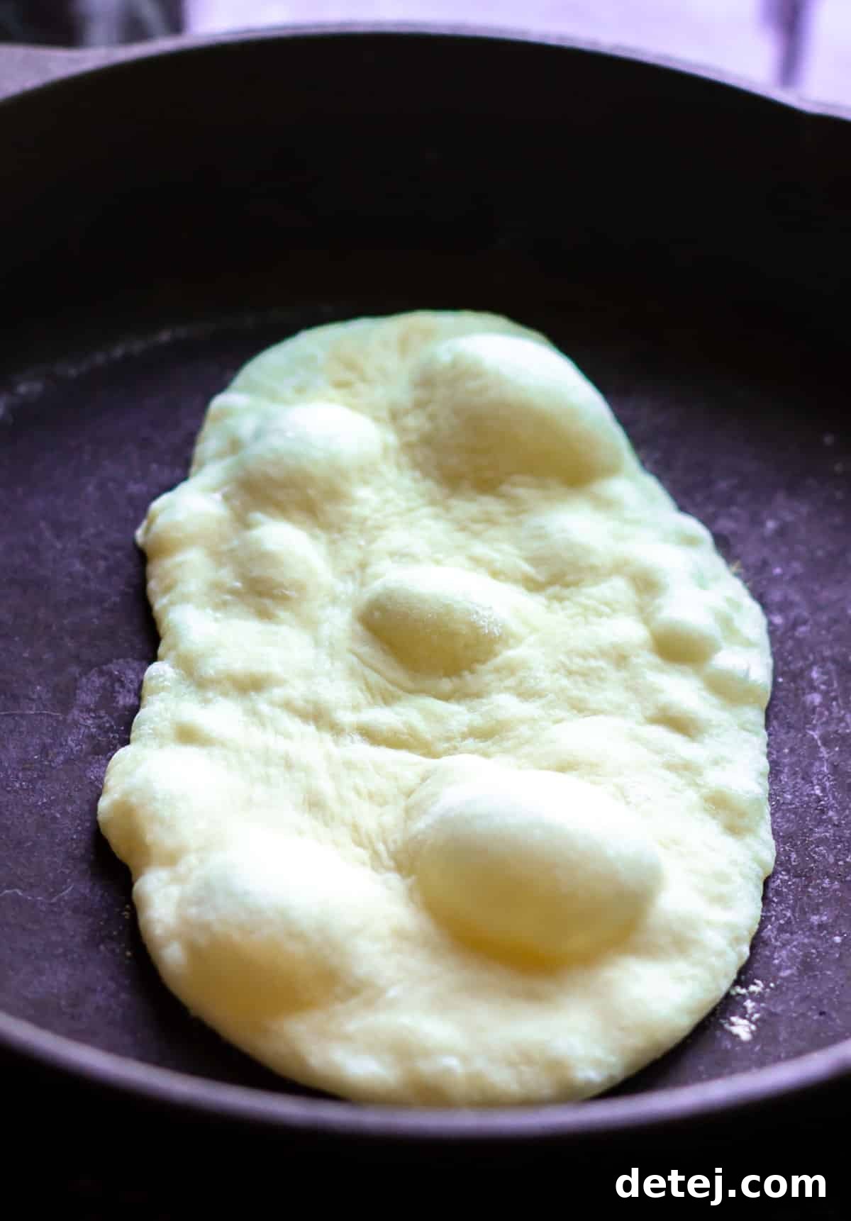 Vegan naan cooking on a hot cast iron pan, showing large bubbles forming on its surface.