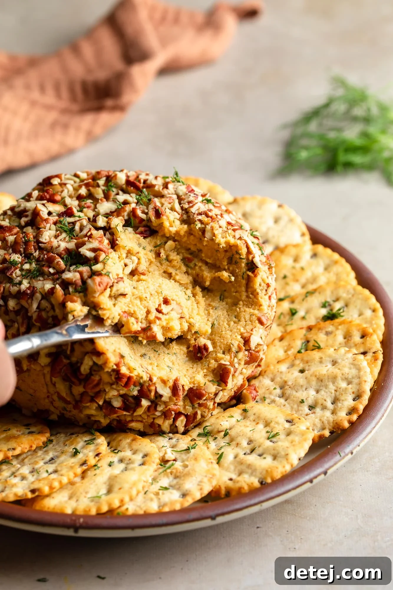 using a knife to lift a scoop from a vegan cheese ball on a plate with crackers.
