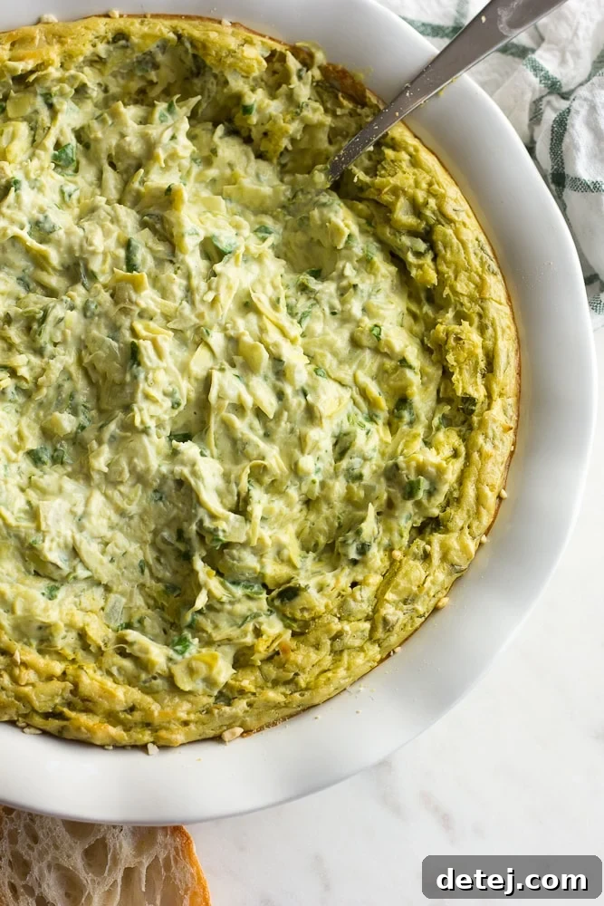 Effortlessly Delicious Vegan Spinach Artichoke Dip 3 A large ceramic bowl filled with creamy vegan spinach artichoke dip, featuring visible pieces of spinach and artichoke, with a serving spoon resting inside.