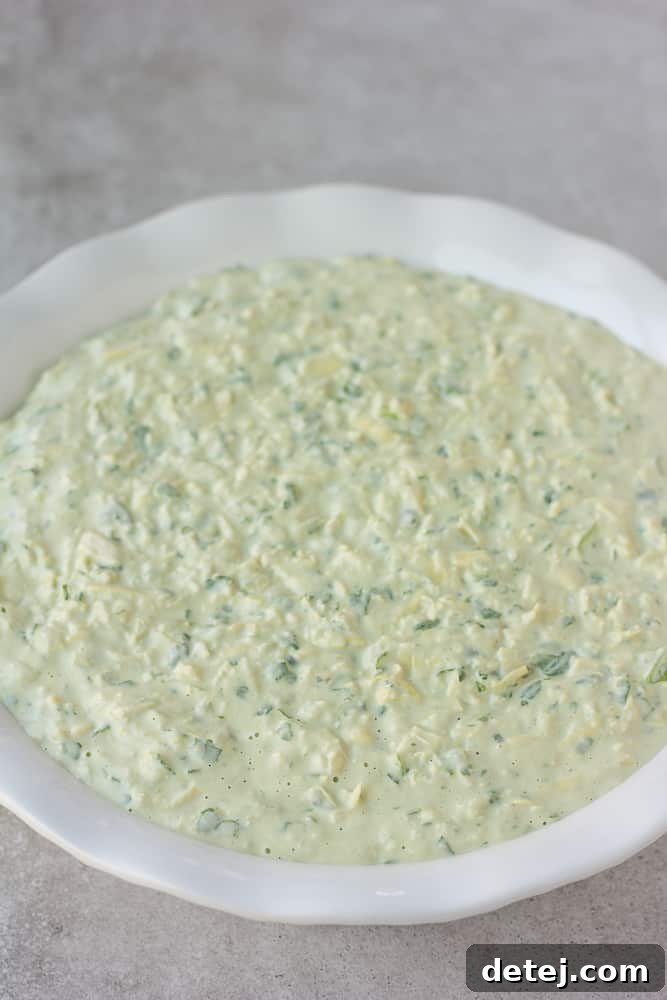 Effortlessly Delicious Vegan Spinach Artichoke Dip 7 An unbaked vegan spinach artichoke dip mixture spread evenly in a round ceramic pie dish, ready to be placed in the oven.