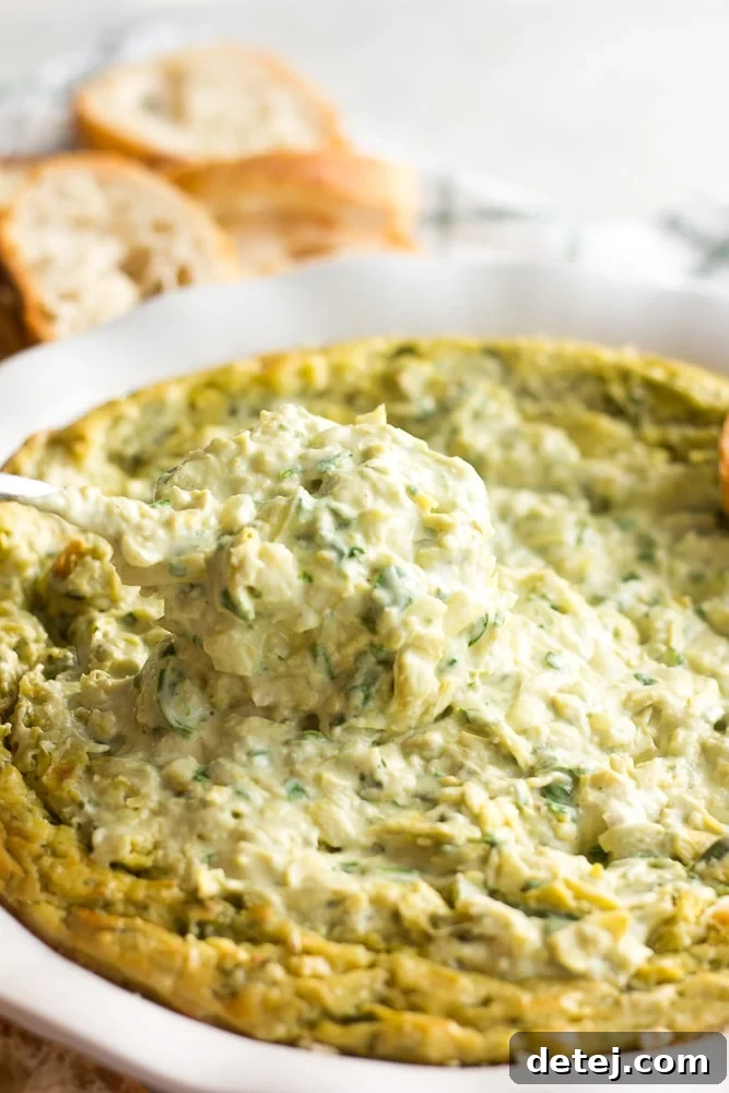 Effortlessly Delicious Vegan Spinach Artichoke Dip 8 A freshly baked vegan spinach artichoke dip, golden and bubbly, with a serving spoon scooping out a portion, highlighting its rich, creamy texture.
