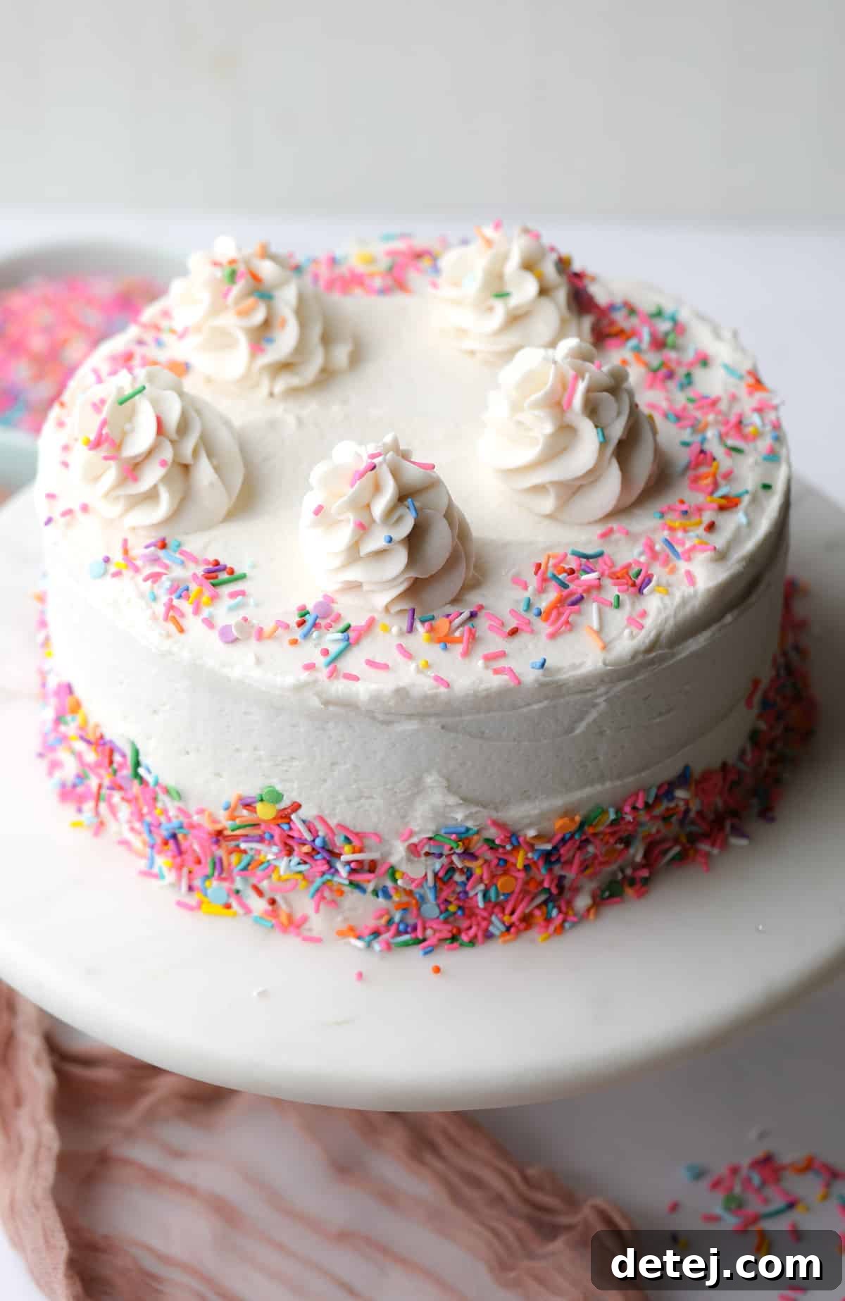 A beautifully decorated and frosted vegan vanilla cake, topped with vibrant colorful sprinkles, set against a clean white background.