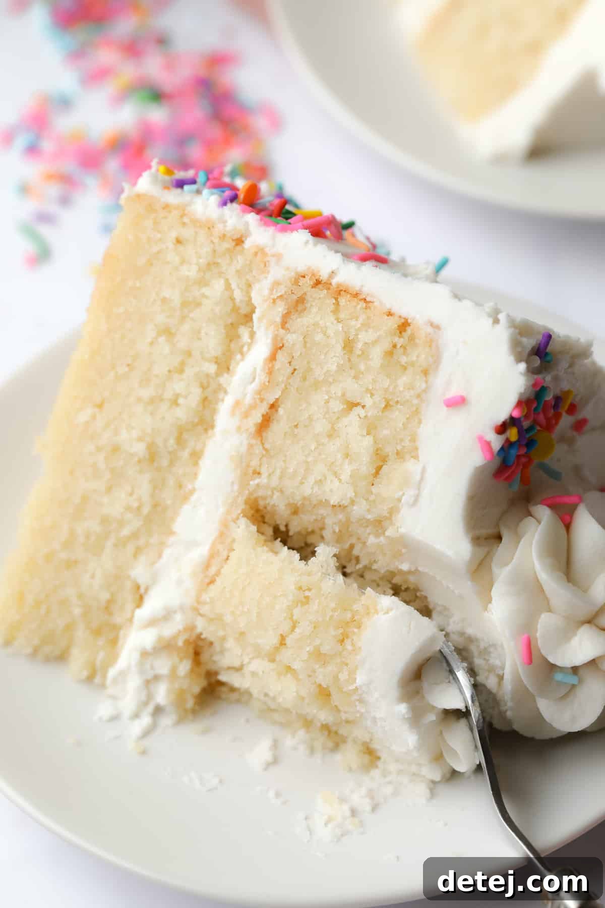 A fork gently scooping a perfect piece of frosted vegan vanilla cake from a white plate, showcasing its soft texture.