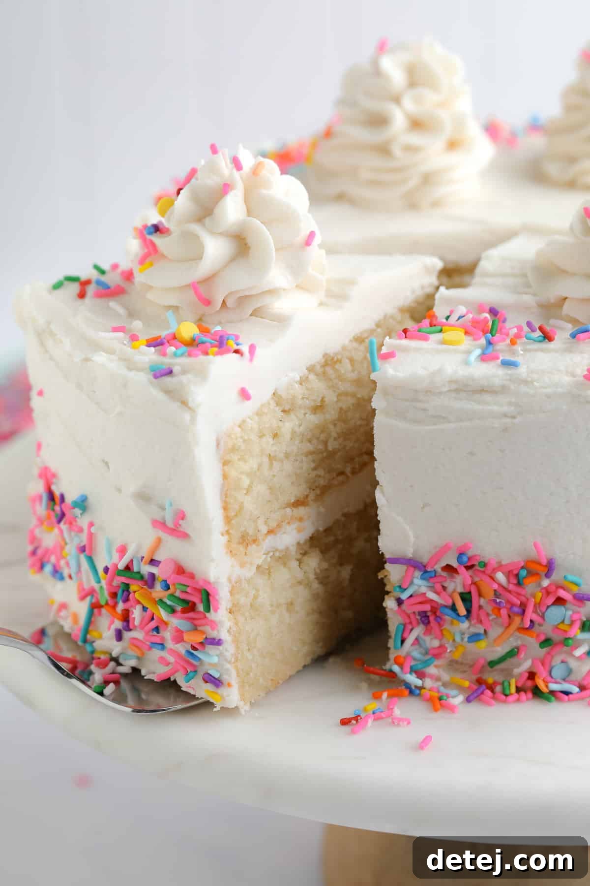 A perfectly baked and frosted vegan vanilla cake on a cake stand, with a serving utensil gracefully lifting a slice, highlighting its moist layers.