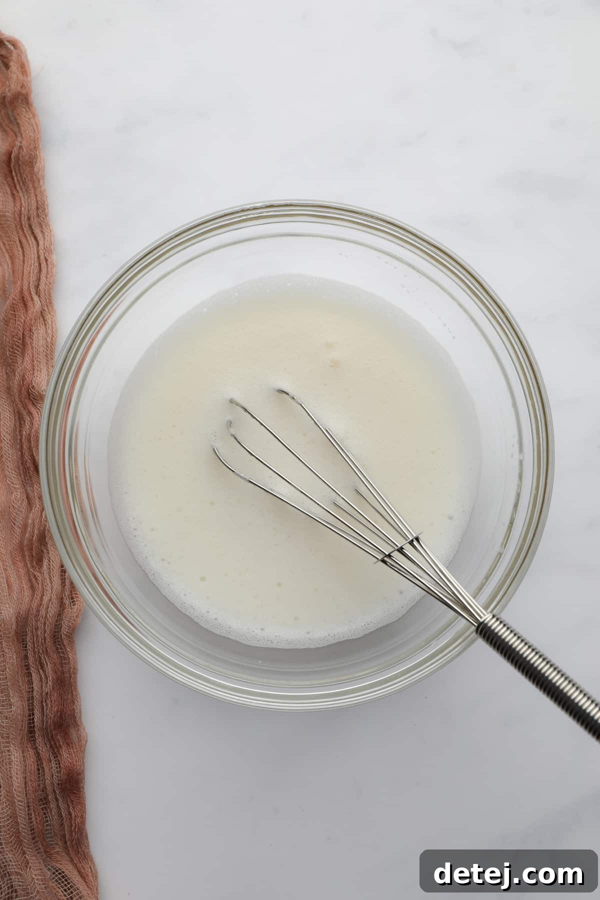 A small bowl of vigorously whisked aquafaba and cream of tartar, showing a light, foamy texture, with a whisk resting inside.