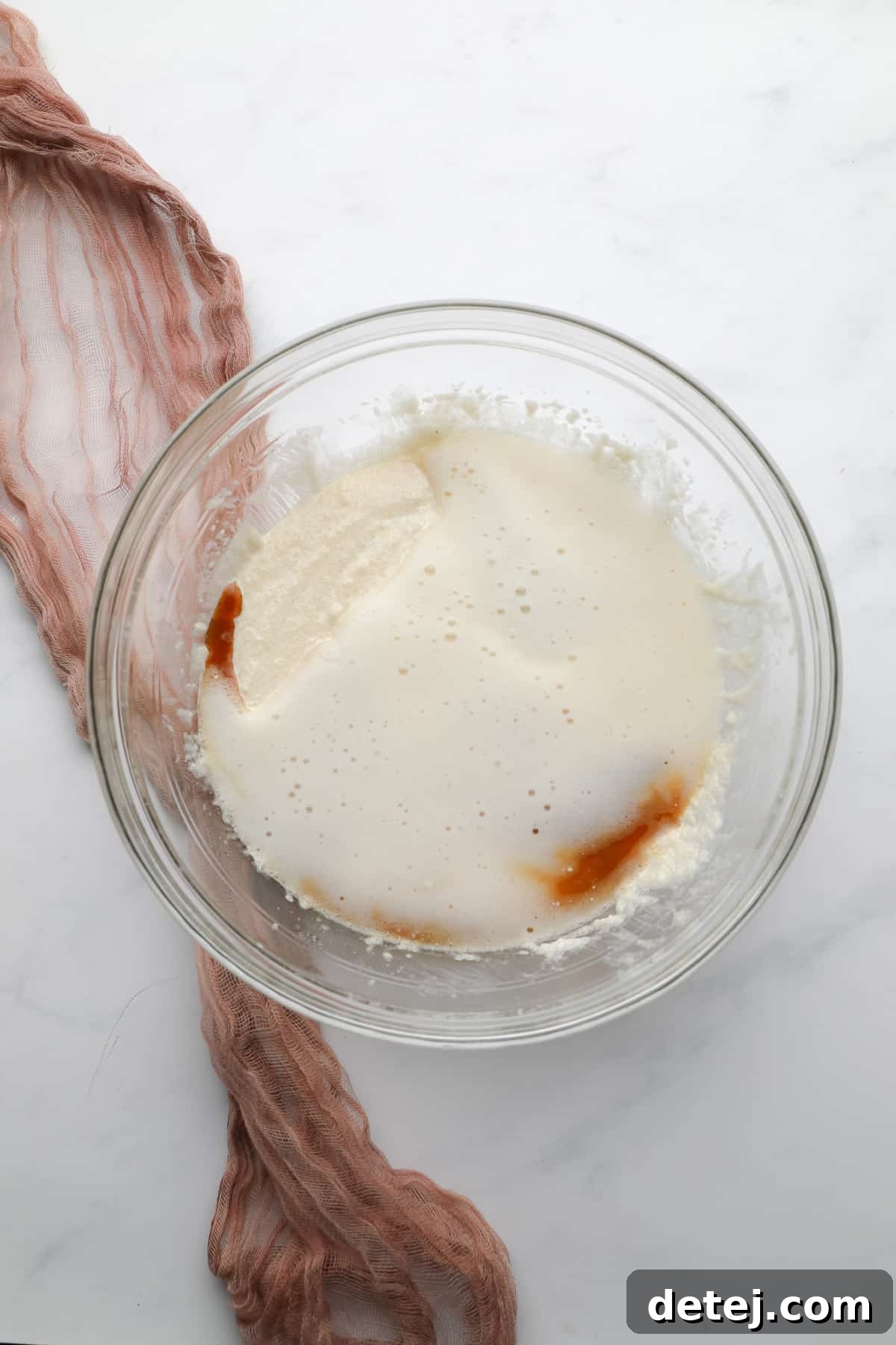 A glass mixing bowl holding a creamy mixture, with vanilla extract and the foamy aquafaba mixture recently added on top, awaiting to be blended.