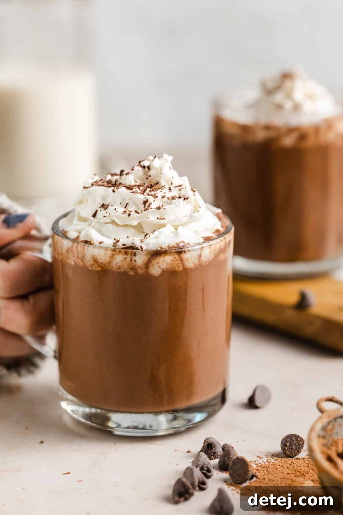 A woman holding a clear glass mug brimming with decadent vegan hot chocolate, beautifully topped with a swirl of plant-based whipped cream and chocolate shavings.