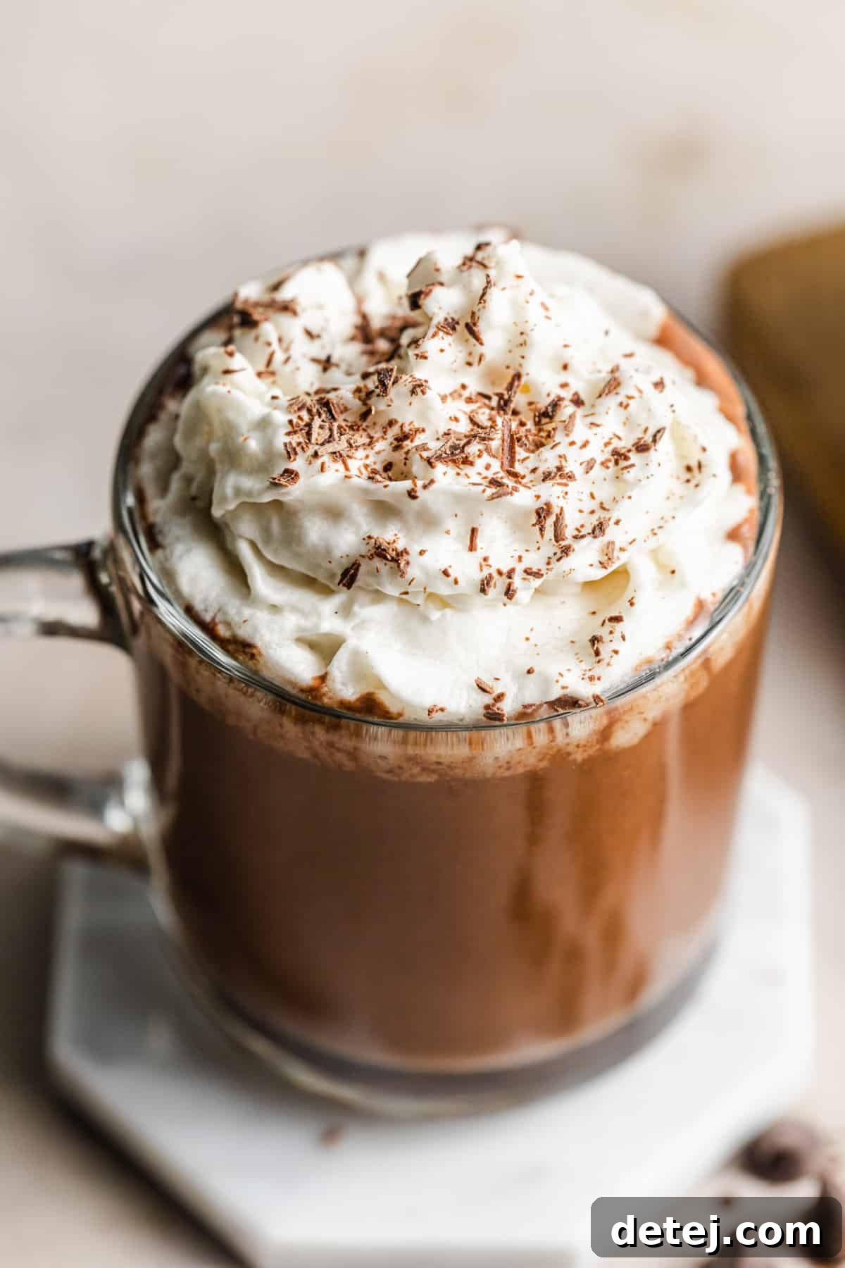 A glass mug filled to the brim with creamy vegan hot chocolate, artfully topped with a generous dollop of whipped cream, resting on an elegant marble plate.