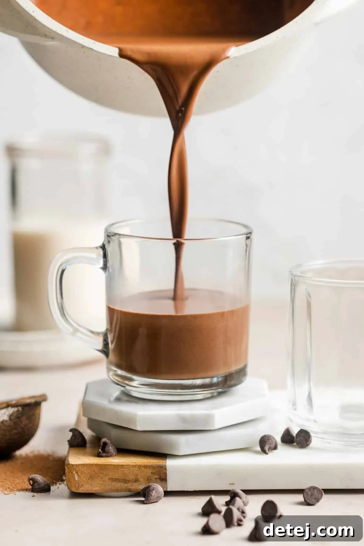 A stream of rich, dark vegan hot chocolate being poured from a white saucepan into a clear glass mug, showcasing its smooth and inviting texture.