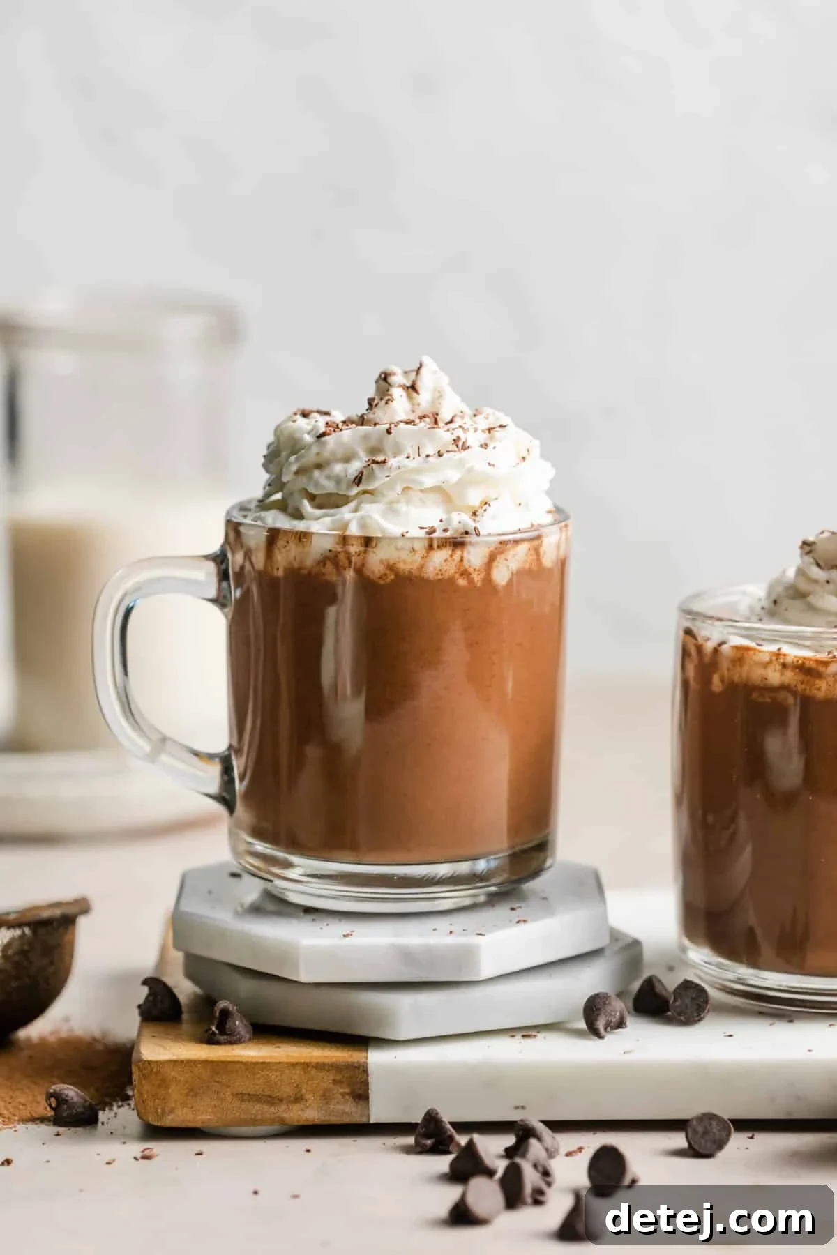 A close-up shot of a glass mug filled with frothy vegan hot chocolate, generously topped with a dollop of fluffy whipped cream and a sprinkle of chocolate shavings.