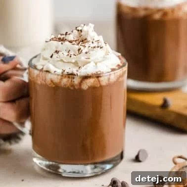 A woman holding onto a glass mug filled with vegan hot chocolate topped with whipped cream.