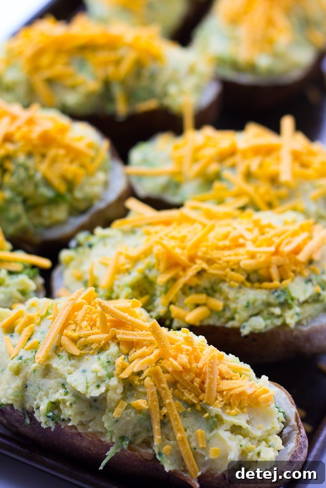 Four potato skins generously filled with the vegan cheesy broccoli mixture, topped with non-dairy cheddar cheese shreds, arranged on a baking sheet, ready for the second bake.