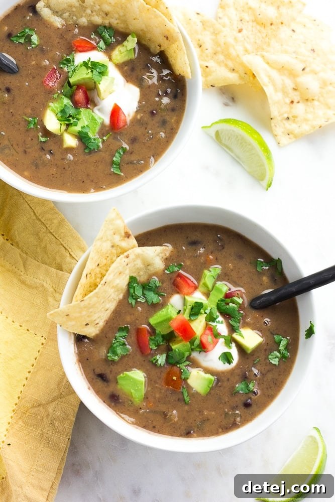 Hearty Vegan Black Bean Soup 2 Two bowls of creamy vegan black bean soup, garnished with avocado, tortilla chips, diced tomatoes, and fresh cilantro, on a wooden table.