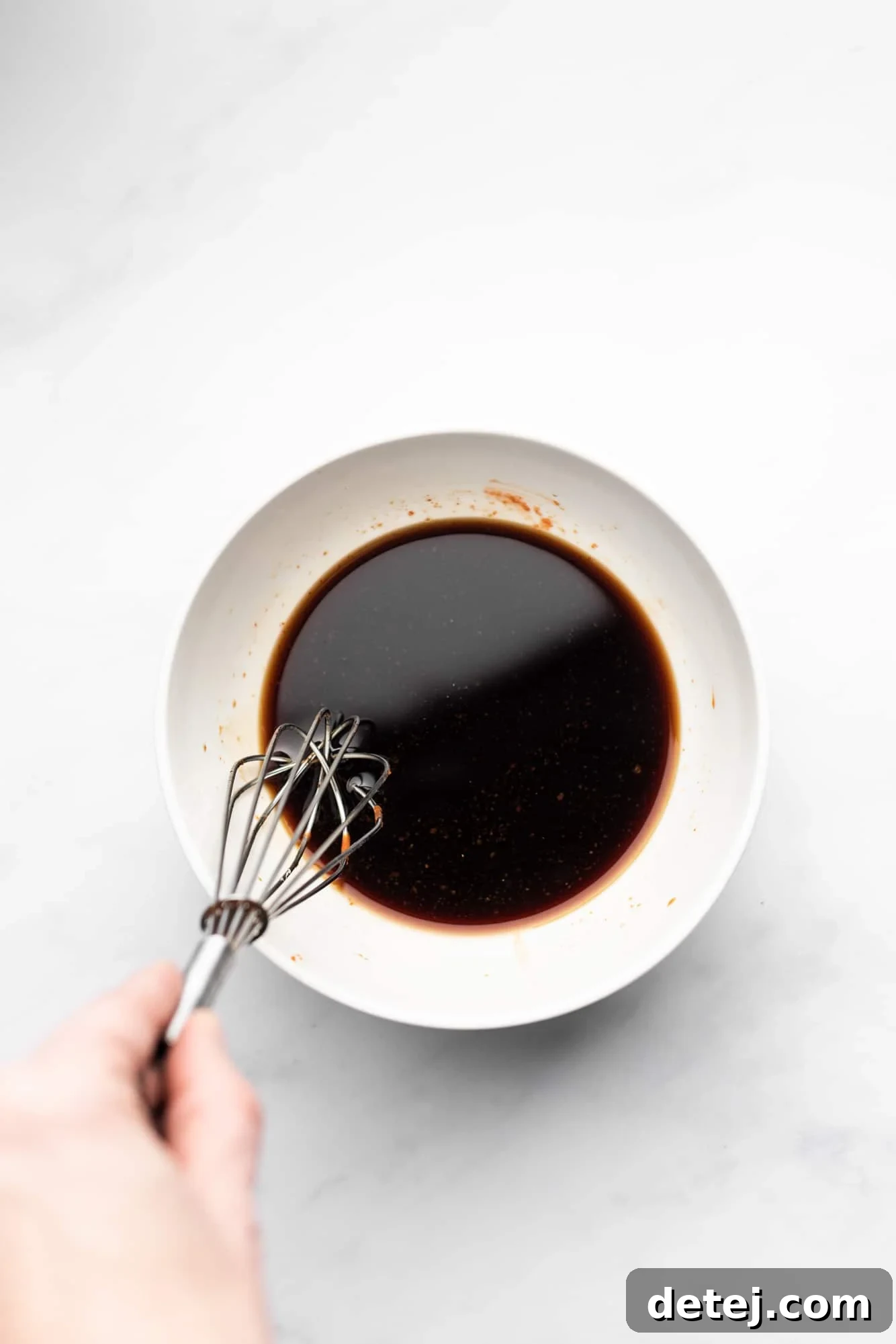 A whisk stirring a rich, dark brown sauce in a clean white bowl, highlighting the sauce preparation step.