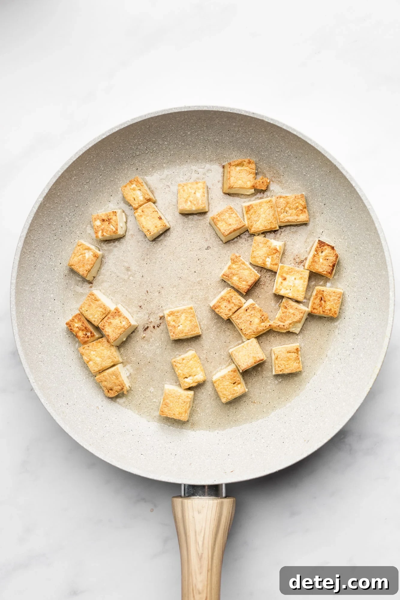 Cubed tofu pieces frying to a golden brown in a beige non-stick pan, showing the cooking process.