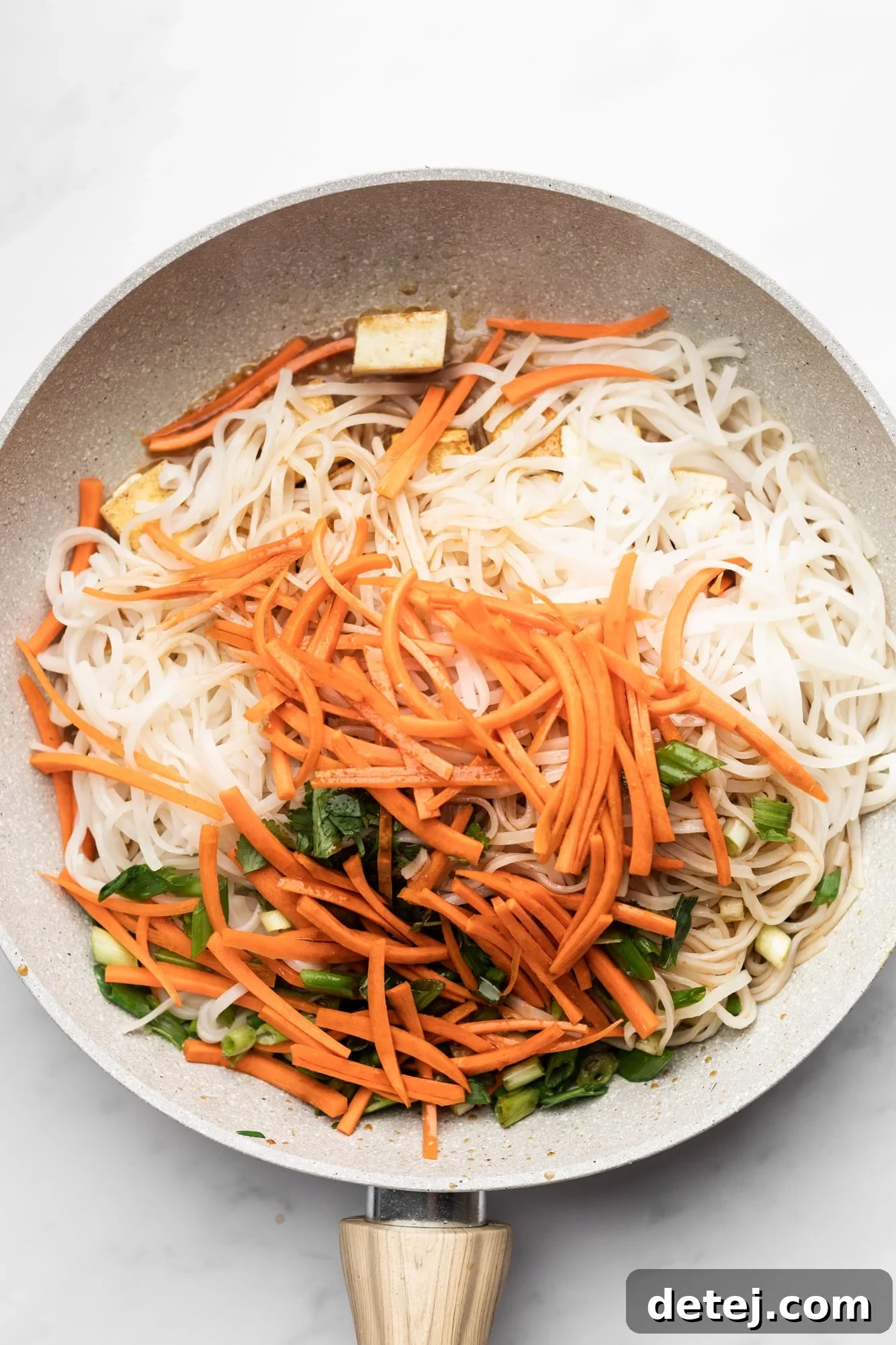 Rice noodles, carrots, and scallions being stirred together with a spoon in a beige pan, demonstrating the final mixing step.