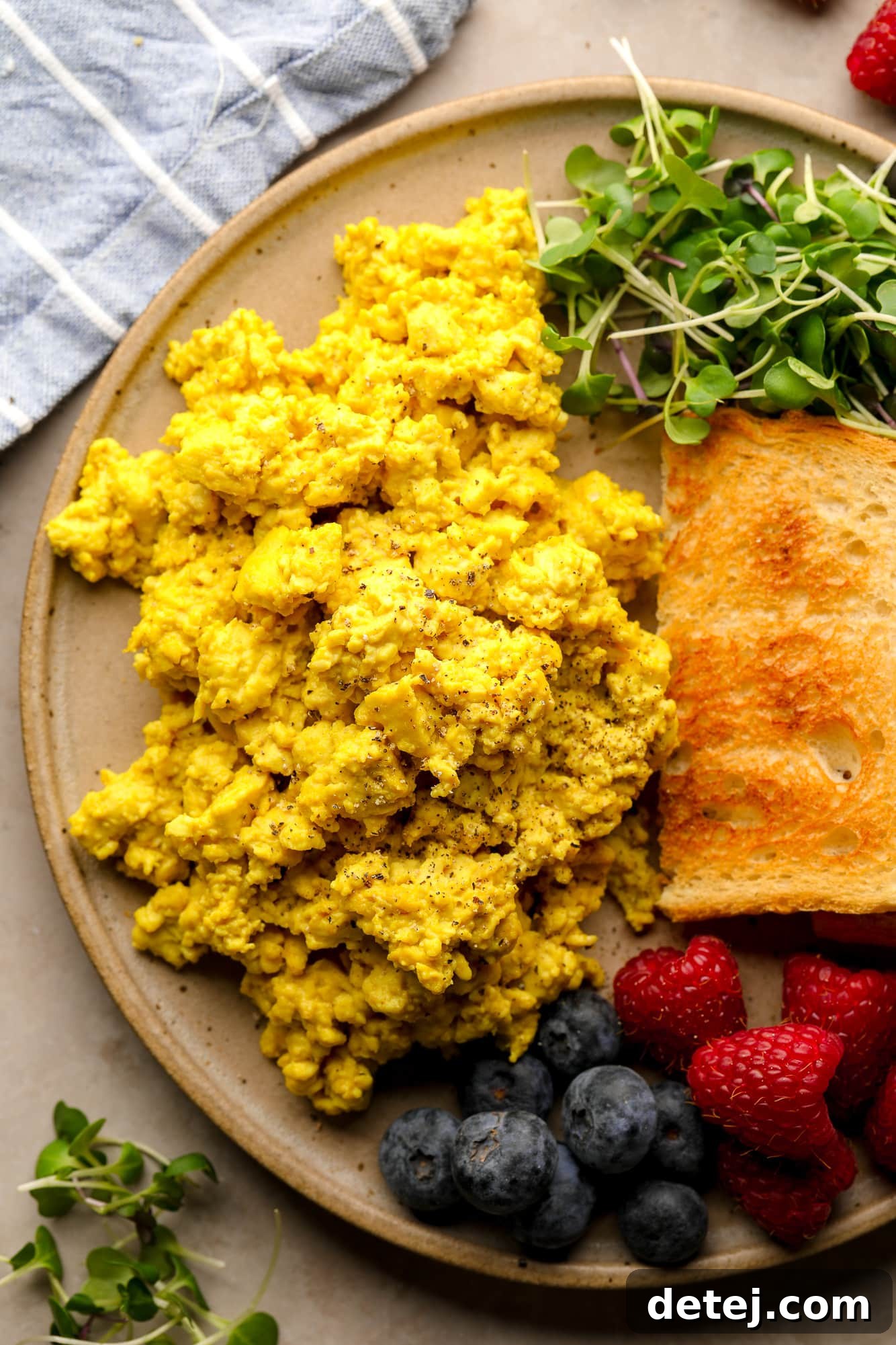 A plate of golden yellow tofu scramble, garnished with fresh herbs, served alongside toasted bread, mixed greens, and fresh berries.