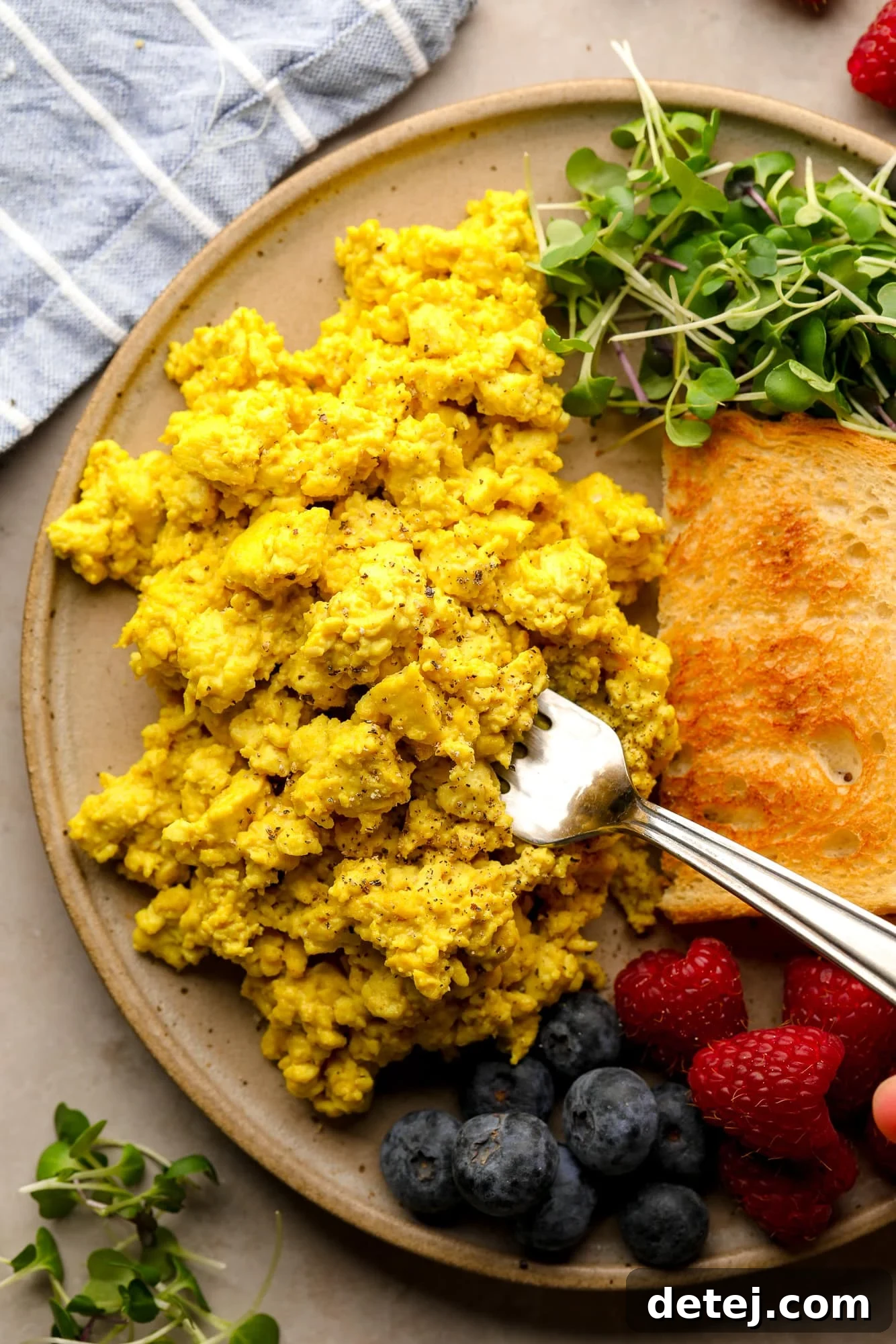 A fork scooping a bite of yellow tofu scramble from a plate, surrounded by toast, fresh greens, and vibrant berries.