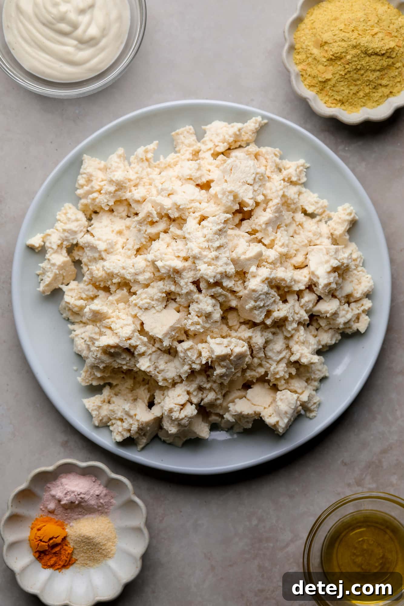 Individual bowls holding each ingredient for tofu scramble: olive oil, a block of tofu, nutritional yeast, black salt, turmeric, garlic powder, and non-dairy milk.