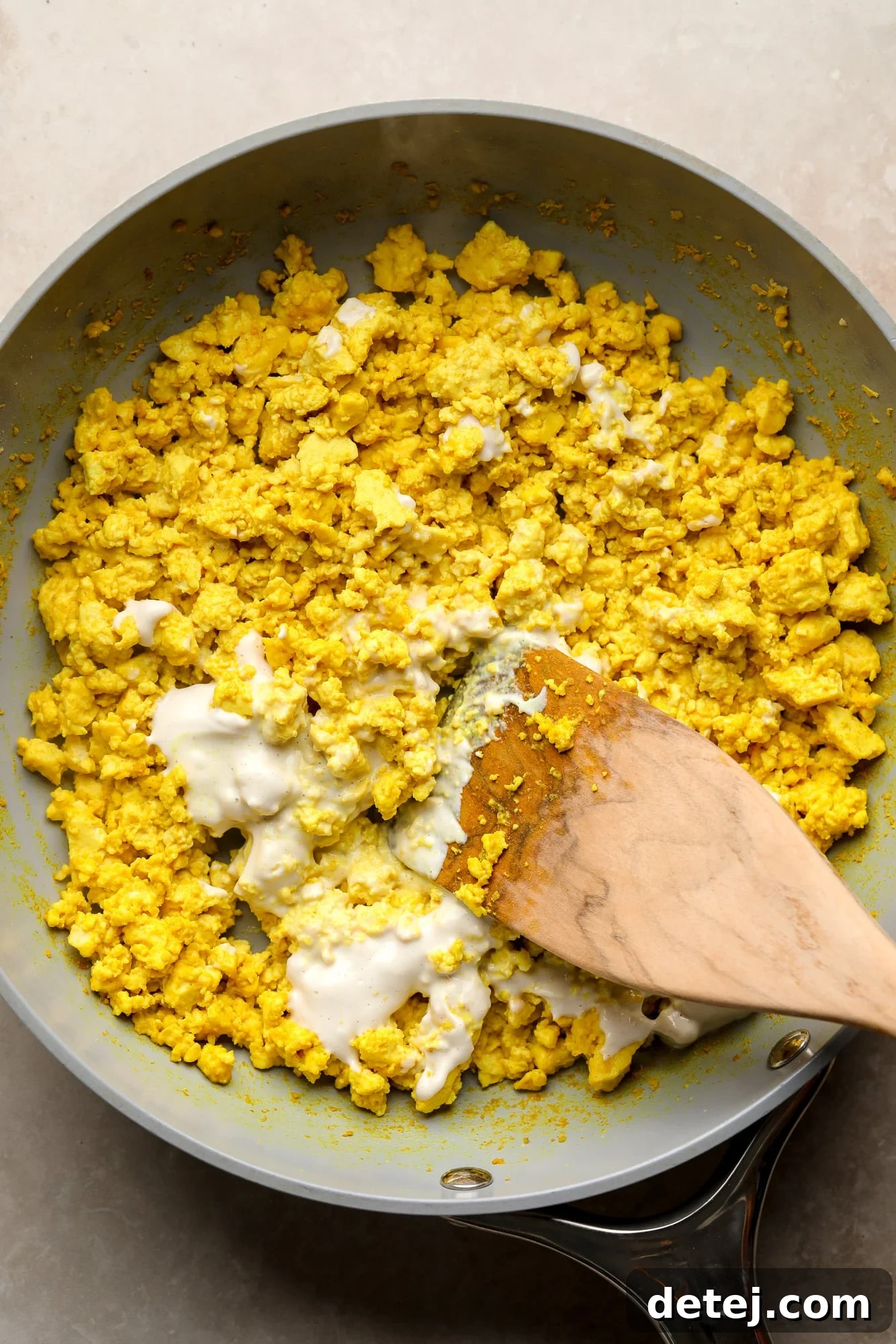 A wooden spoon stirring a splash of non-dairy milk into a batch of yellow tofu scramble in a skillet.