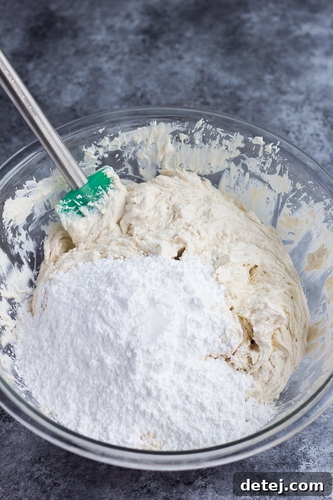 Powdered sugar being carefully added into a bowl of vegan cream cheese frosting, showing the progression of thickening the mixture.
