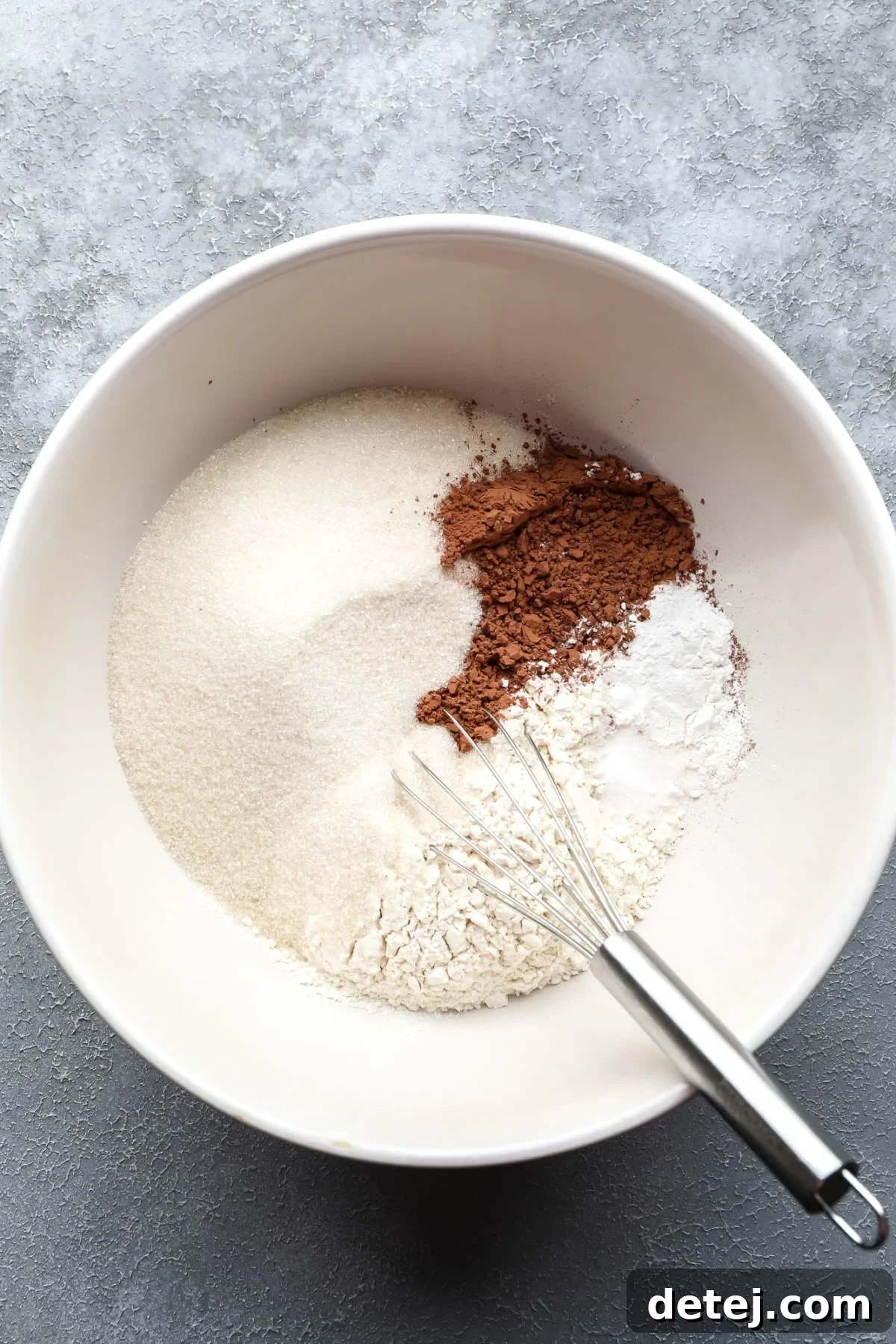 Ruby Red Vegan Indulgence 4 A bowl filled with dry ingredients like flour, sugar, and cocoa powder, with a whisk resting inside, ready for mixing on a grey background.