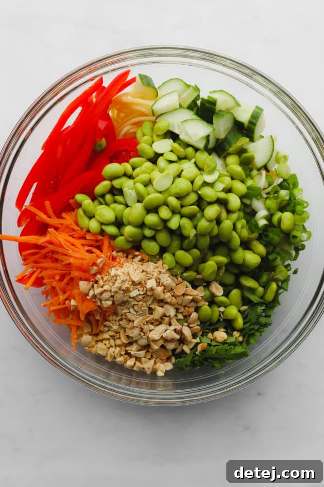 A vibrant glass bowl showcasing an assortment of ingredients: shelled edamame, julienned red pepper, shredded carrots, crushed peanuts, fresh cilantro, sliced green onions, and crisp cucumber slices, all ready for the Thai peanut noodle dish.