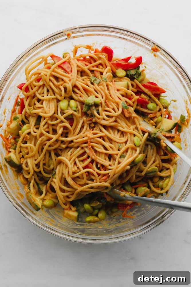 Tongs stirring a medley of noodles, crisp vegetables, and rich peanut sauce in a large glass bowl, illustrating the combining step of the recipe.