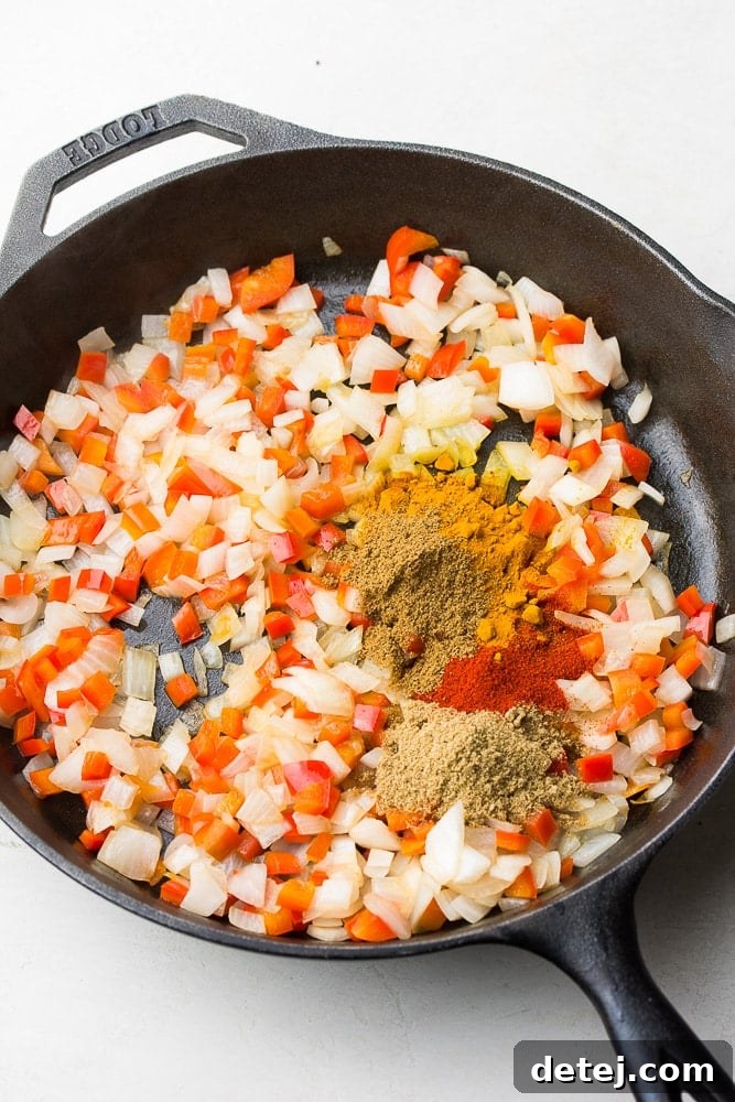 A blend of Indian spices being carefully added to a sautéing mixture of peppers and onions in a hot pan, enhancing the aromatic base.