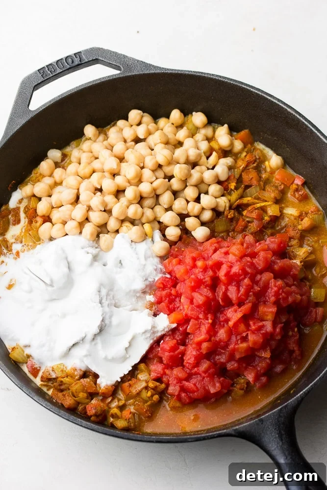 A close-up of a pan containing rich coconut milk, plump chickpeas, and diced tomatoes, layered before being fully mixed into a vibrant curry.