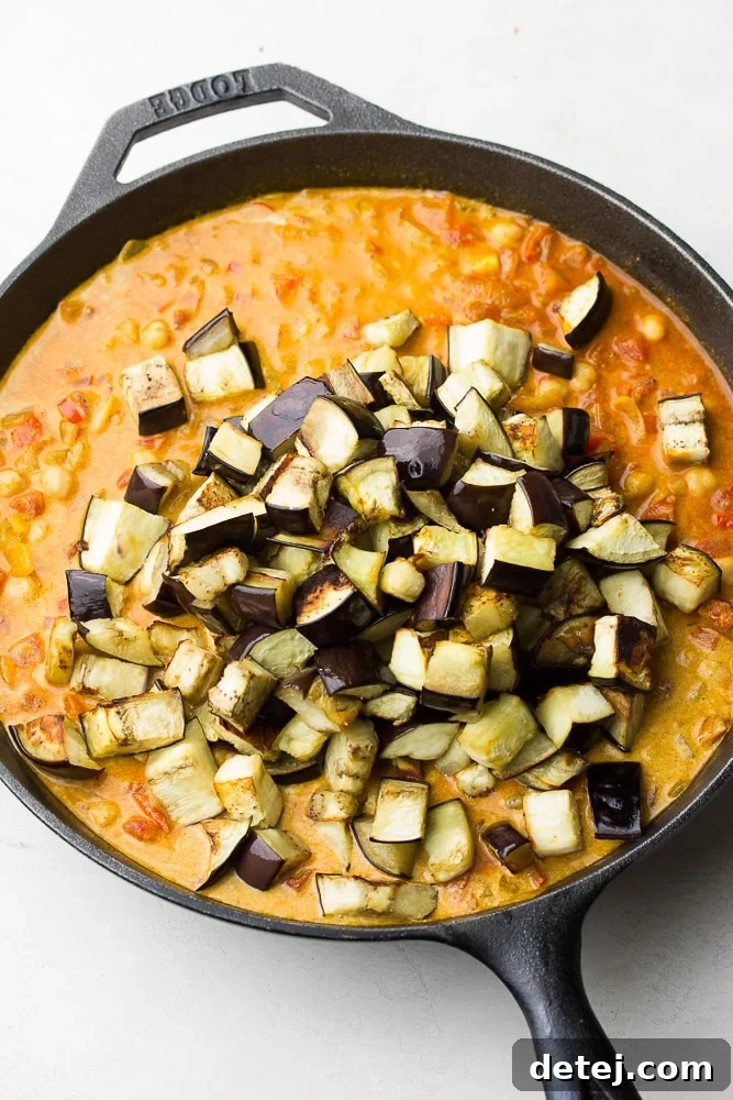 Beautifully roasted eggplant chunks being gently folded into a simmering chickpea and tomato curry sauce in a large skillet, ready to combine flavors.