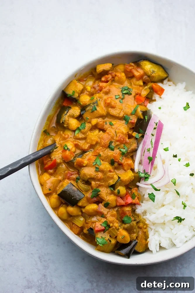 A comforting bowl of roasted eggplant and chickpea curry, served with fluffy white rice and garnished with fresh green herbs.