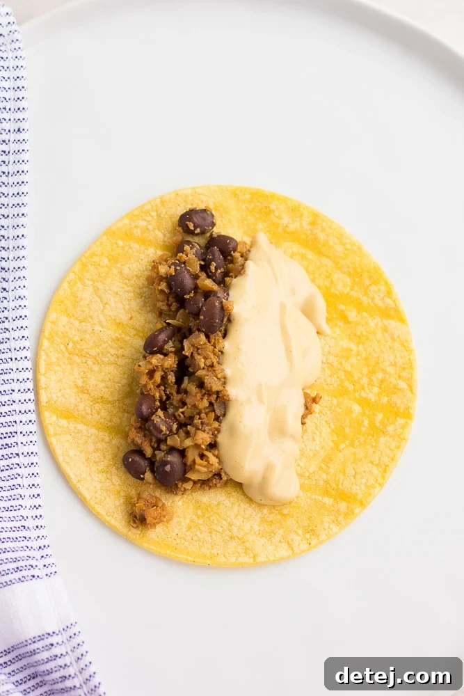 Ultimate Vegan Enchiladas 7 A small corn tortilla filled with savory bean mixture and creamy cheese sauce, resting on a white plate, ready to be rolled.