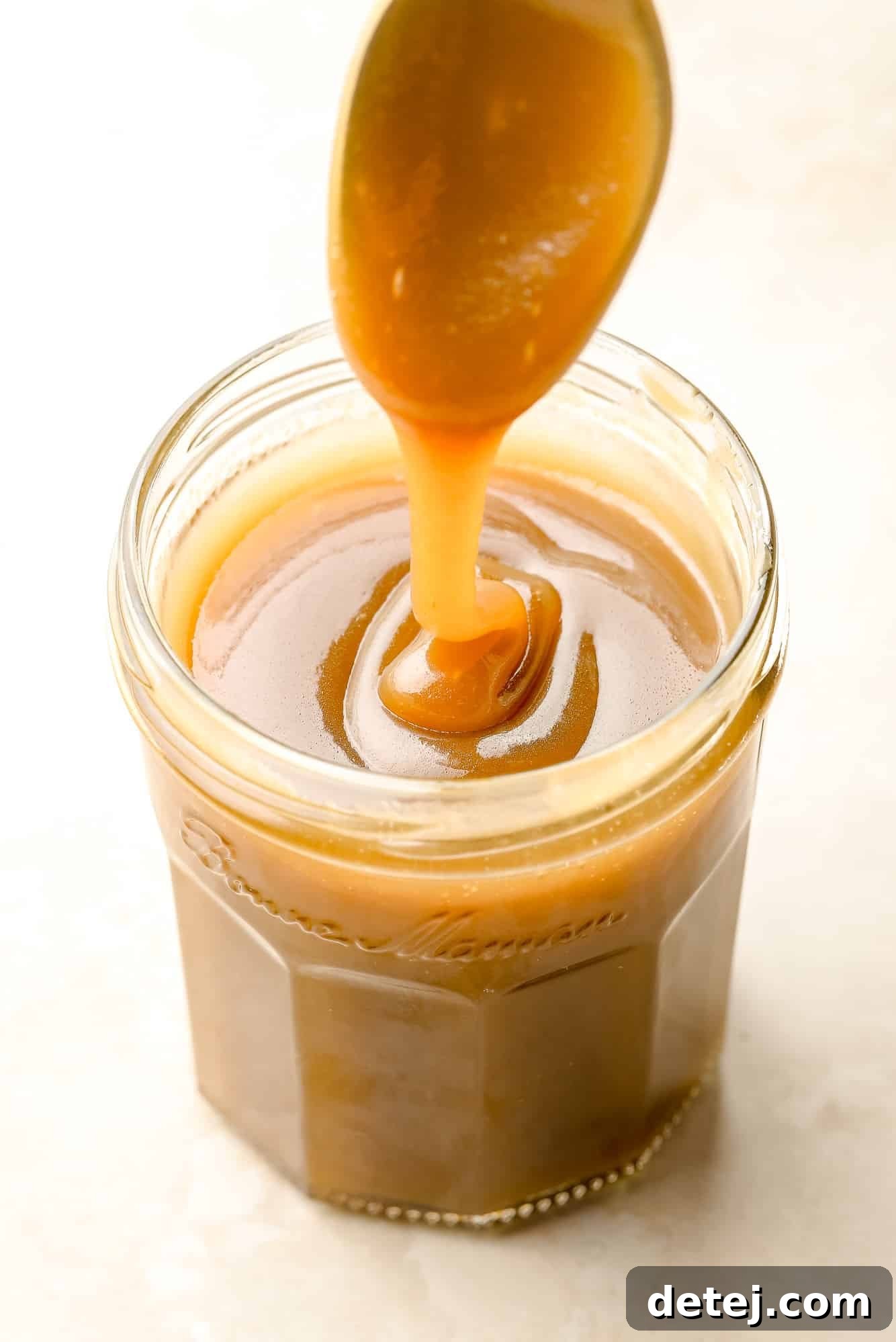 A spoon lifting a stream of silky smooth vegan caramel sauce from a clear glass jar.