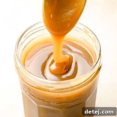 A spoon lifting a stream of silky smooth vegan caramel sauce from a clear glass jar.
