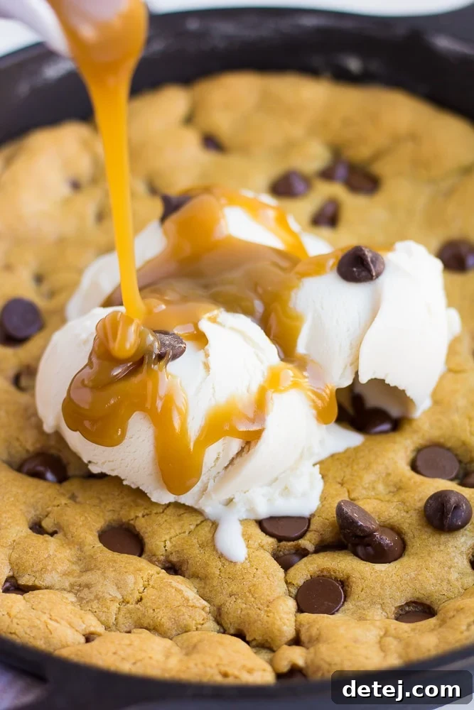 A stream of rich vegan salted caramel sauce being poured over a warm skillet cookie topped with scoops of non-dairy vanilla ice cream.