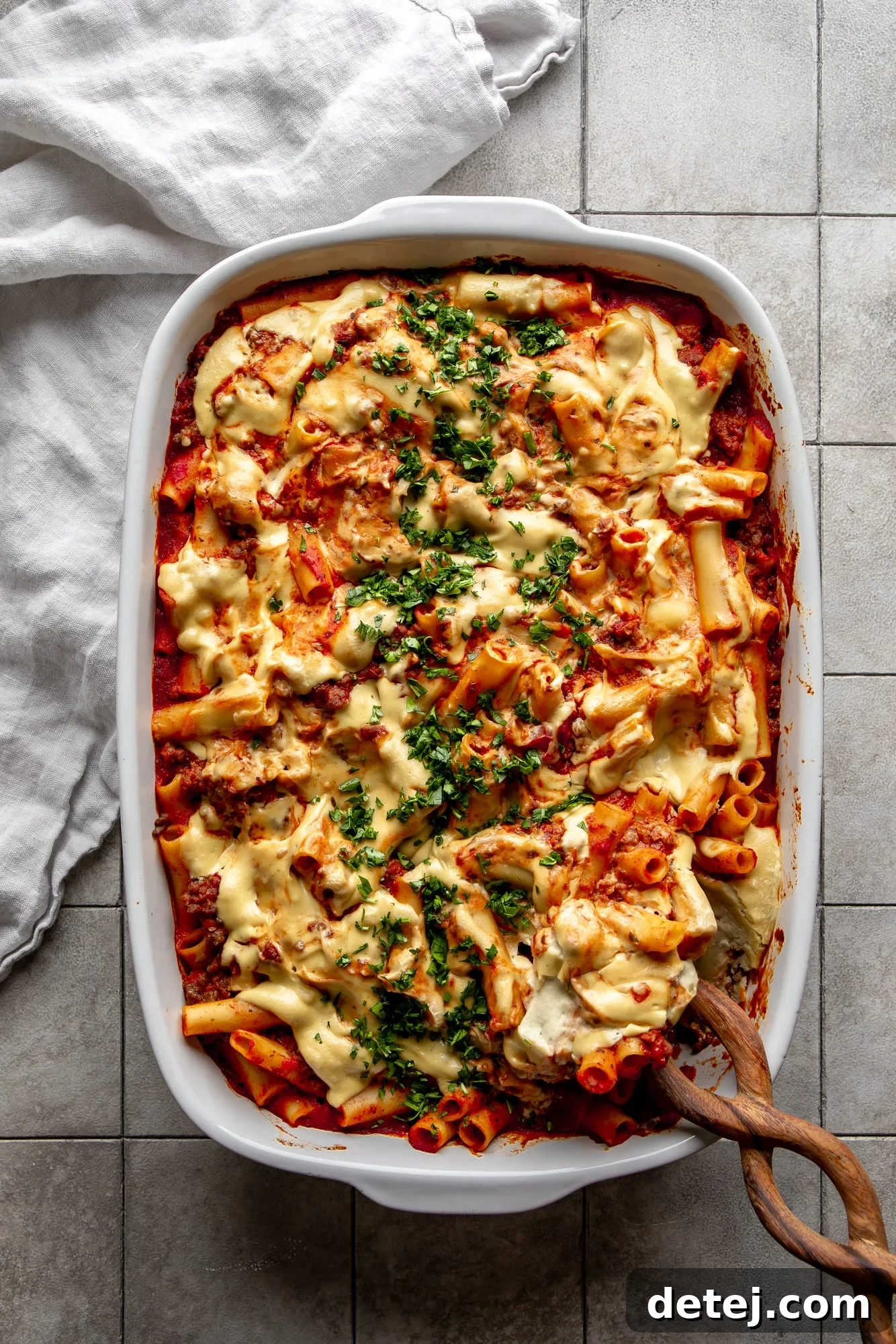 A wooden spoon carefully scooping a generous portion of Vegan Baked Ziti from a bubbling casserole dish, revealing the creamy layers of pasta and cheese.