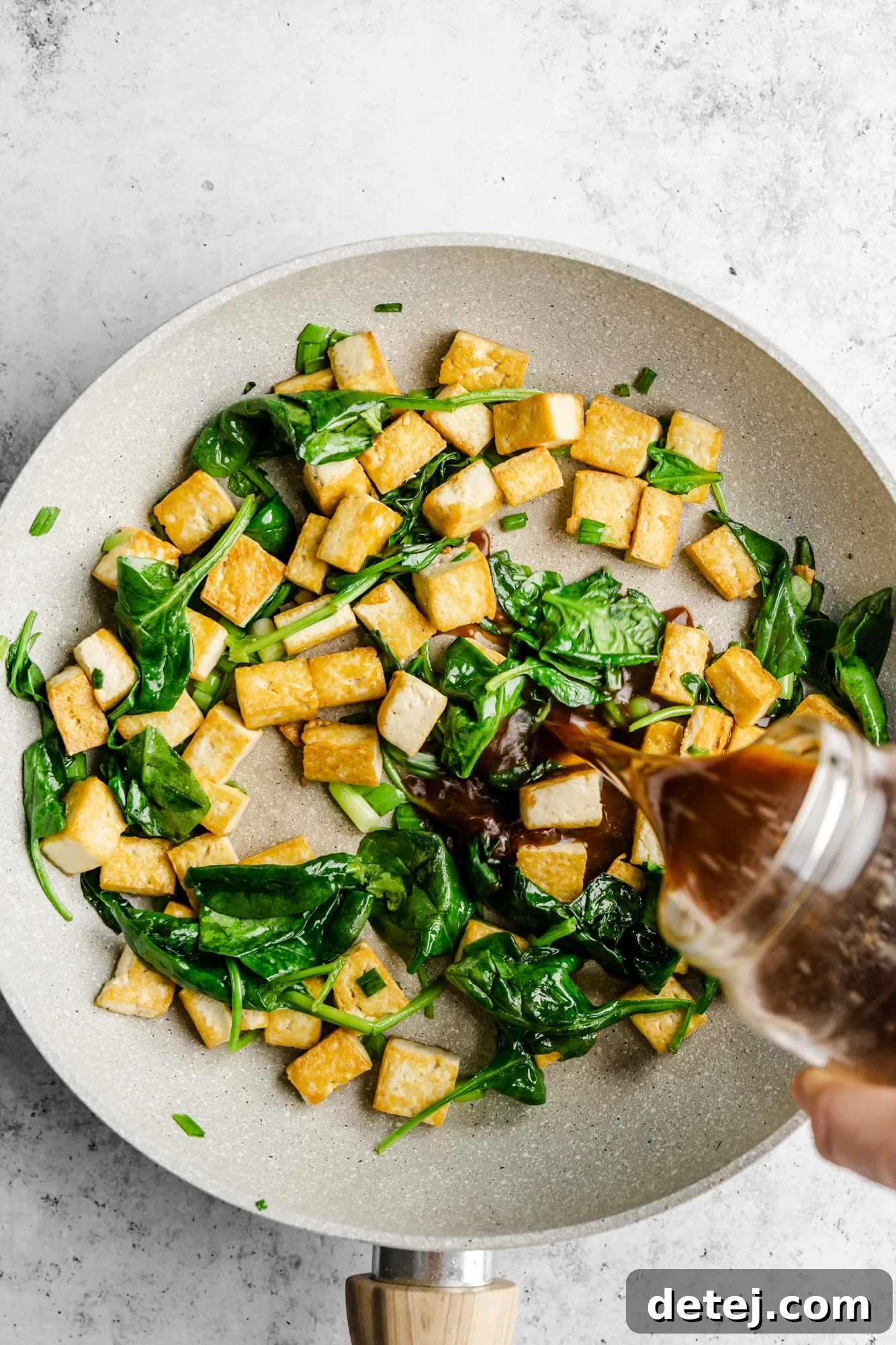 Effortless One Pan Tofu Stir Fry 6 Pouring stir fry sauce in a grey skillet with tofu and spinach.