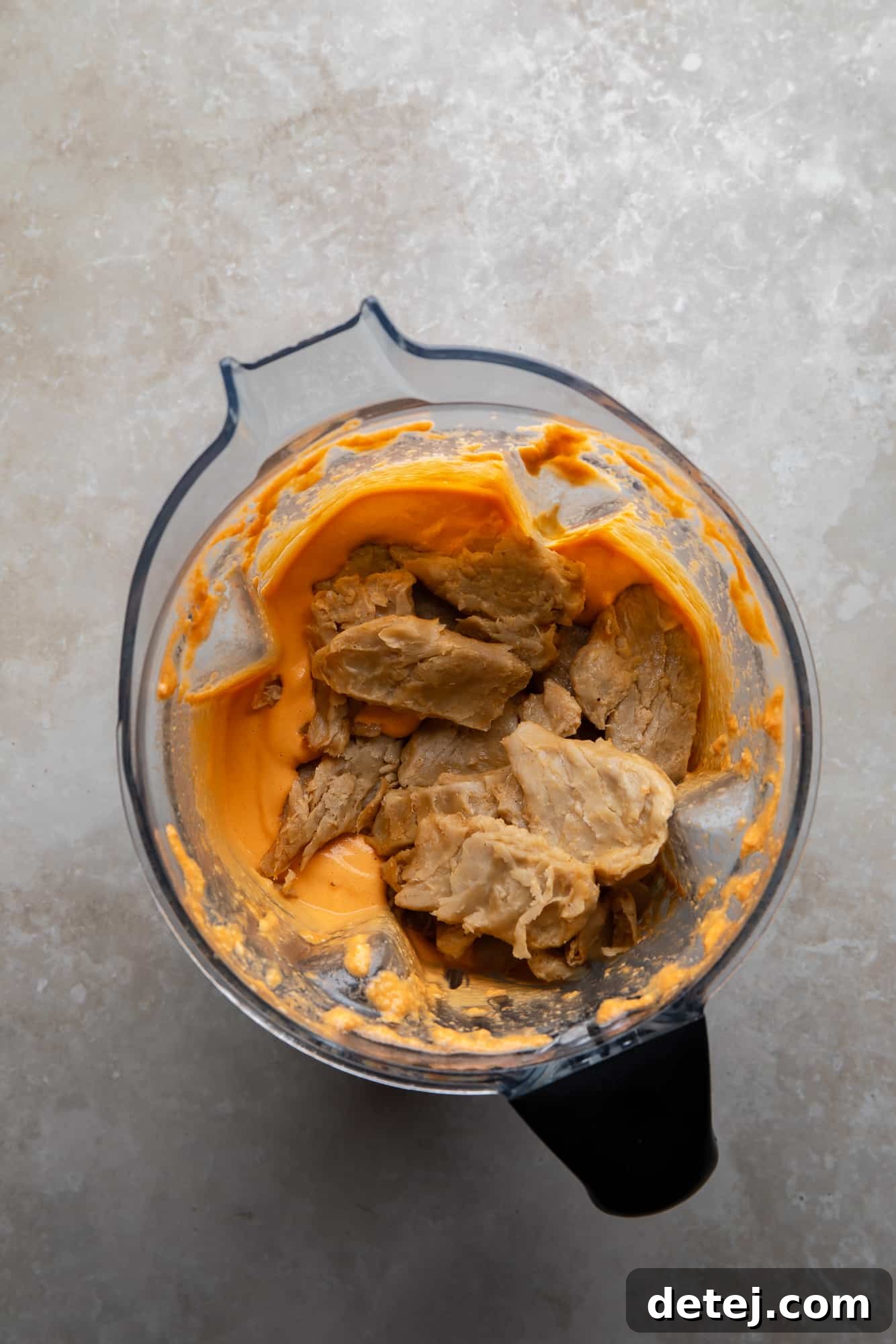 Vegan chicken pieces being added to the blended vegan buffalo chicken dip in a large blender.