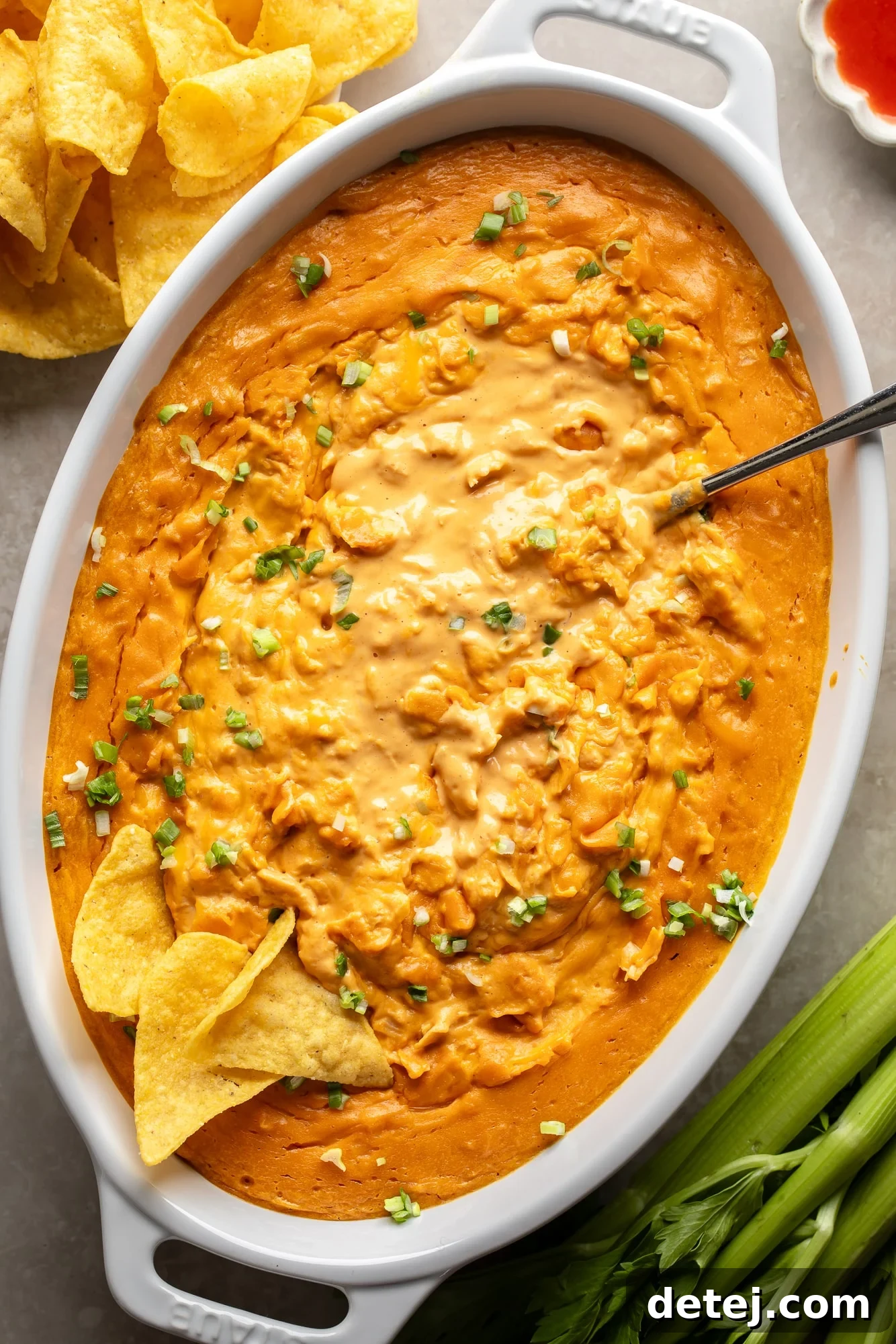 Overhead view of vegan buffalo chicken dip in a large white baking dish, garnished with green onions and surrounded by tortilla chips, ready to be devoured.