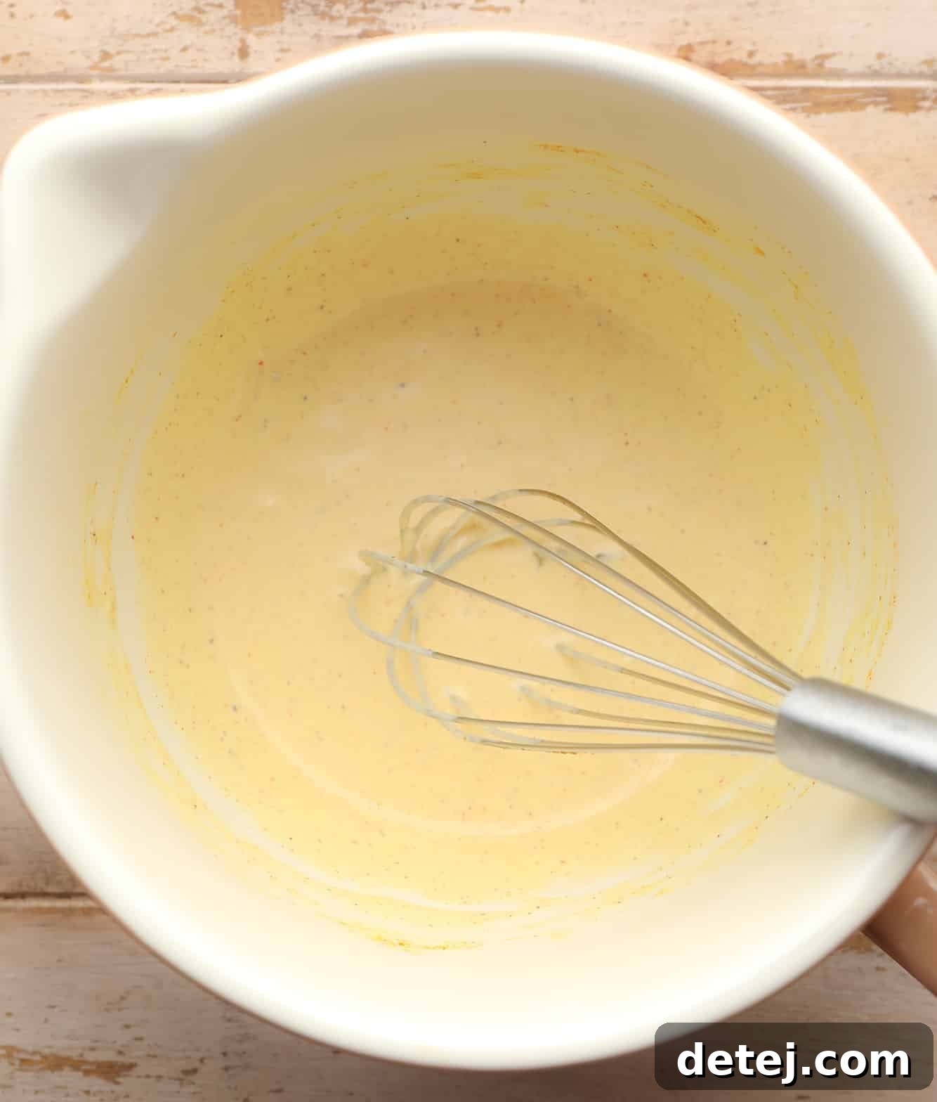 a whisk being used to create a creamy yellow dressing in a white bowl, showing the preparation of the vegan egg salad base.