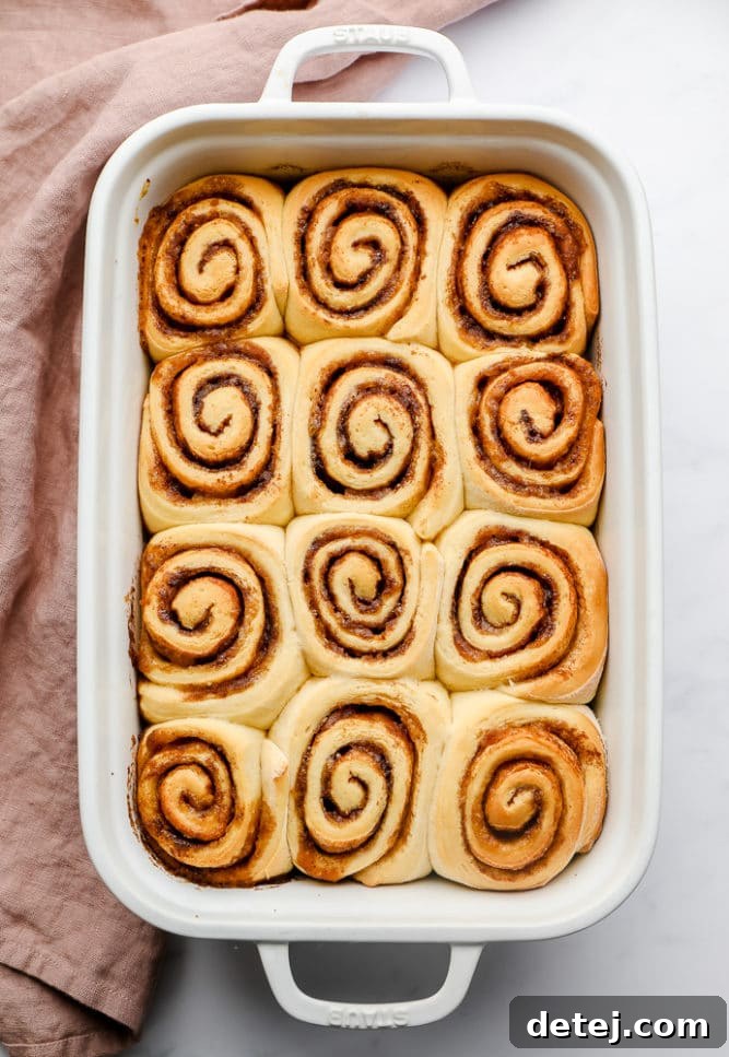 Cooked, golden brown cinnamon rolls in a baking dish, with a pink towel nearby, awaiting frosting.