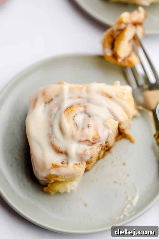 A single vegan cinnamon roll on a grey plate, with a fork taking a bite, showing the gooey center.