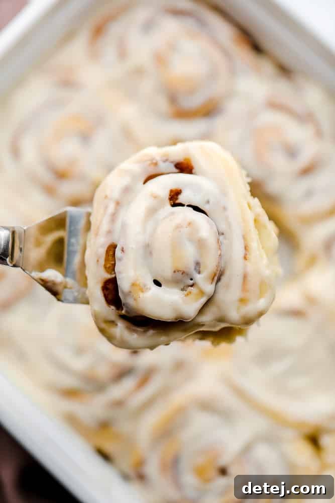 A spatula lifting a perfect vegan cinnamon roll, revealing more frosted rolls in a baking dish below.